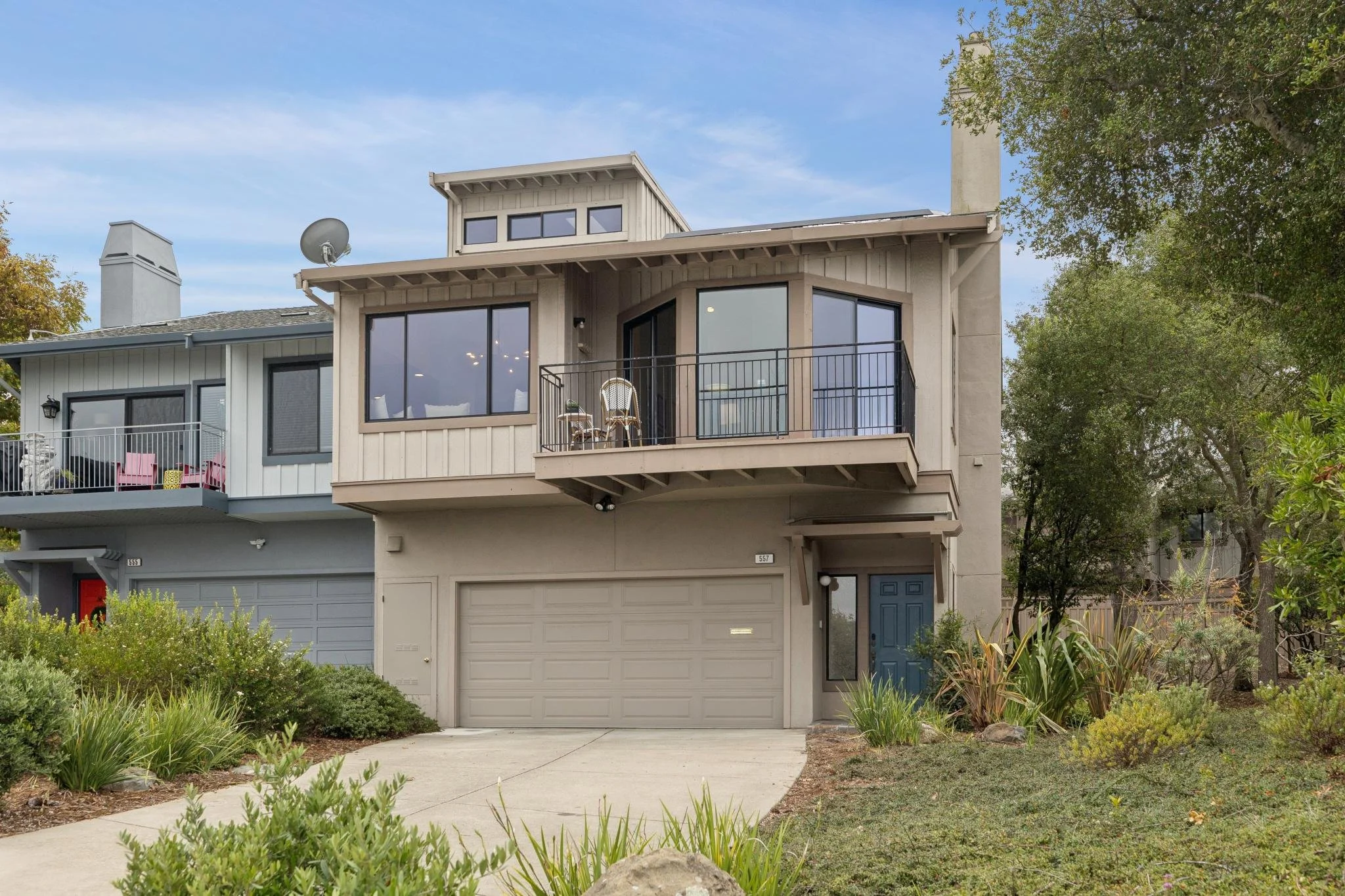 Three-story modern house with a garage on the ground level, a balcony with chairs, large windows, and a small garden in front, surrounded by trees and greenery.