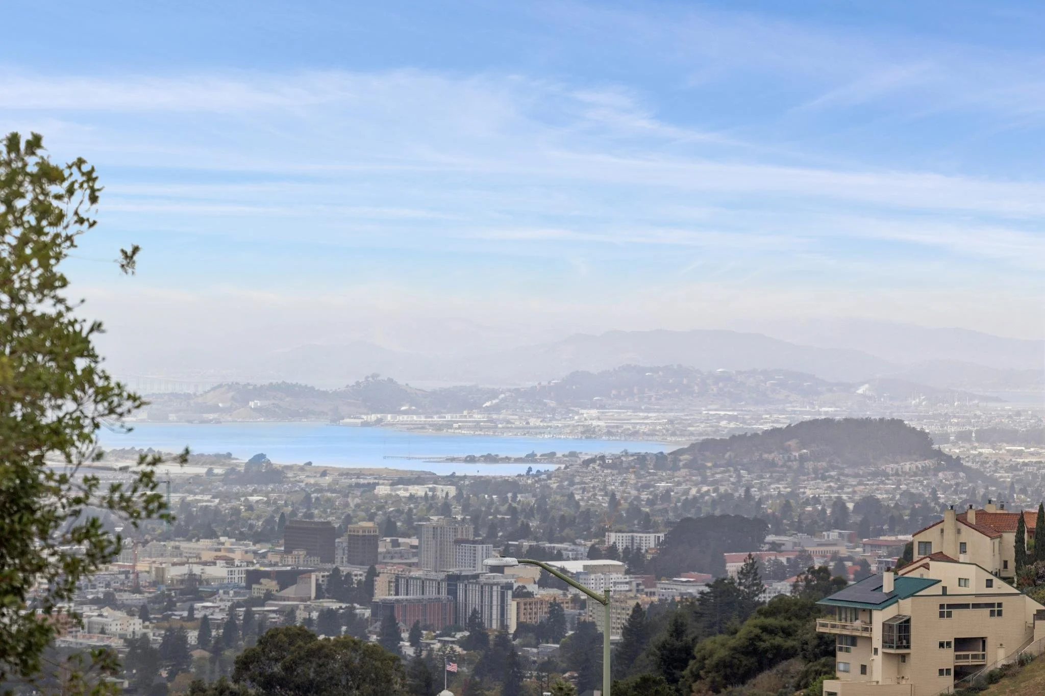 Panoramic view of a cityscape with various buildings and a distant bay, under a partly cloudy sky.