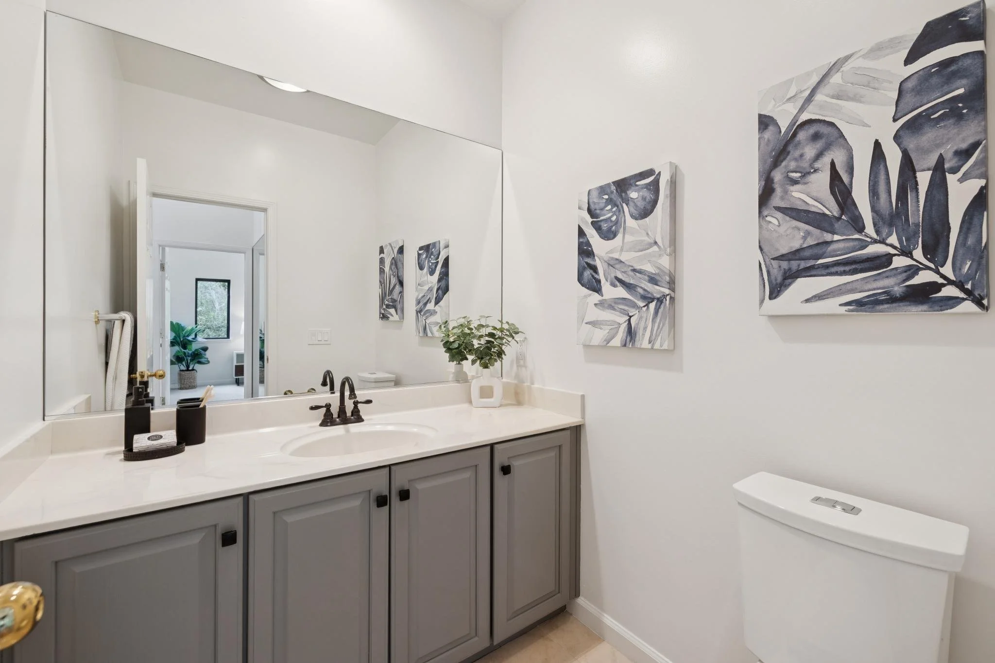 A bathroom featuring a white countertop with a sink, a large mirror, artwork with dark botanical prints on the wall, and a glimpse into a room with a window and potted plant through the doorway.