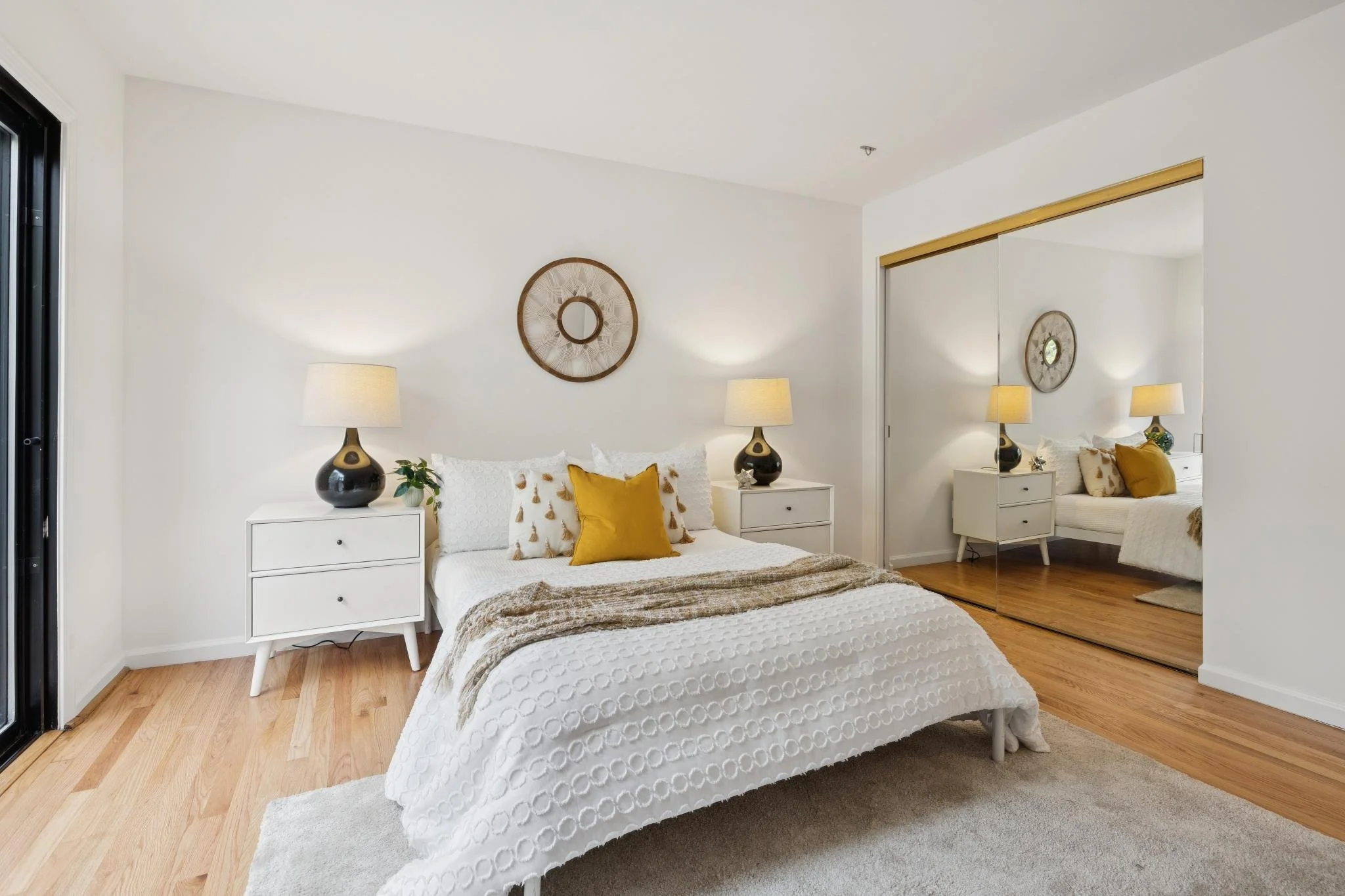 A bedroom with a white bedspread, a yellow pillow, and a brown blanket. There are two white nightstands with black lamps, a potted plant, and wall decor with a round mirror on the wall. A large mirror closet door reflects the room.