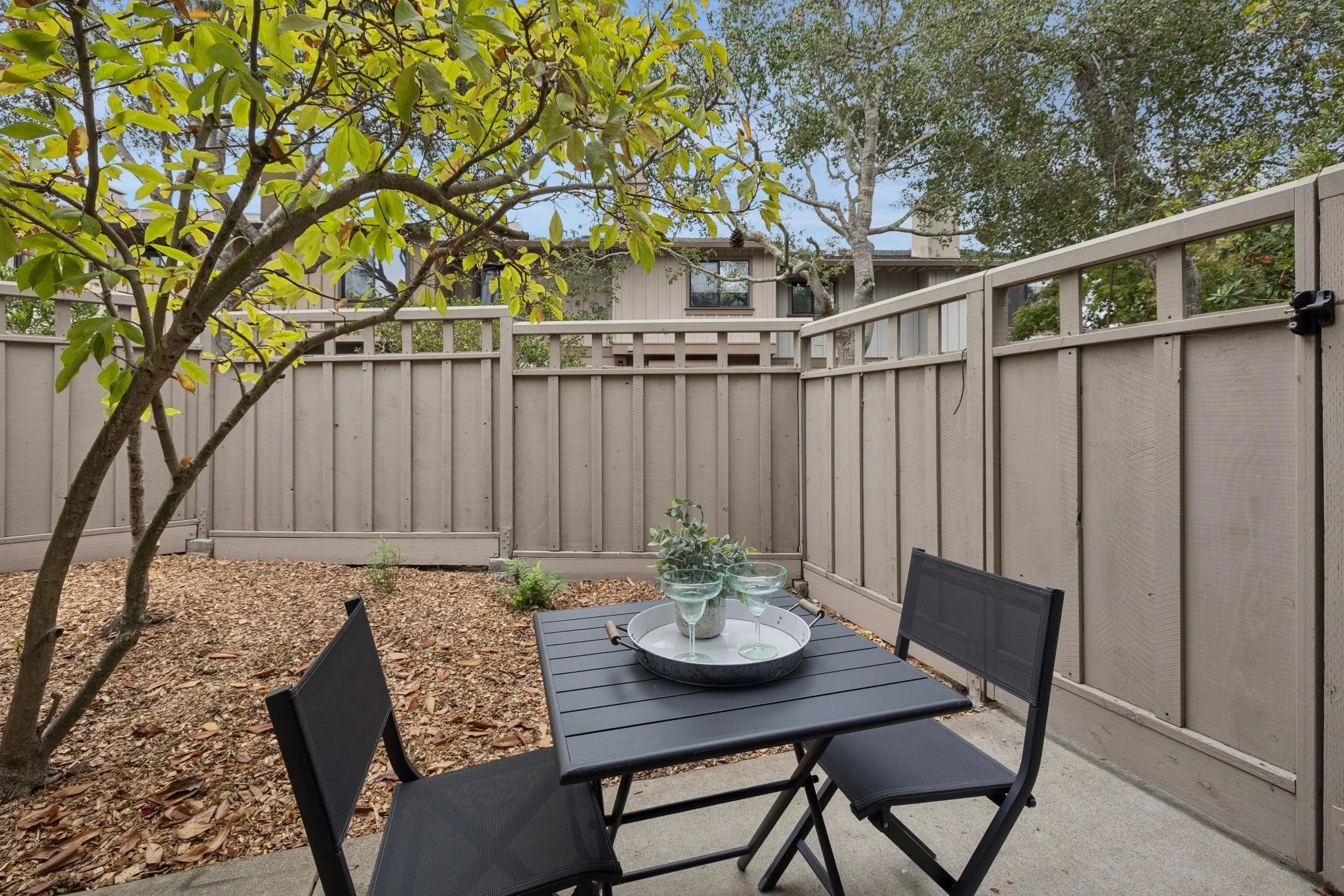 A backyard patio with a black table and four matching chairs, a tray with glasses, a tree with green leaves, a beige wooden fence, and a house with windows in the background.