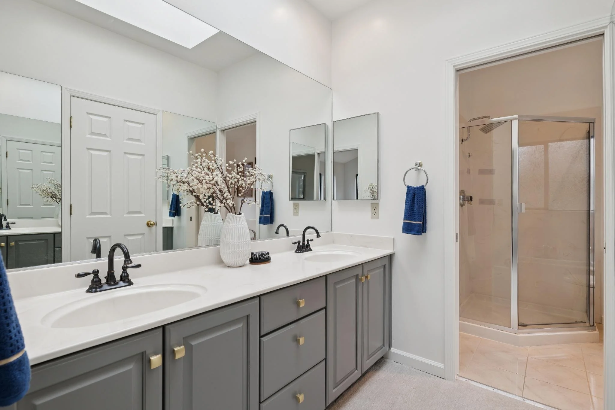 A modern bathroom with a double sink vanity with gray cabinets, white countertop, two black faucets, and a large wall mirror. There is a white decorative vase with branches on the counter. Blue towels hang on the wall and a glass shower with a chrome