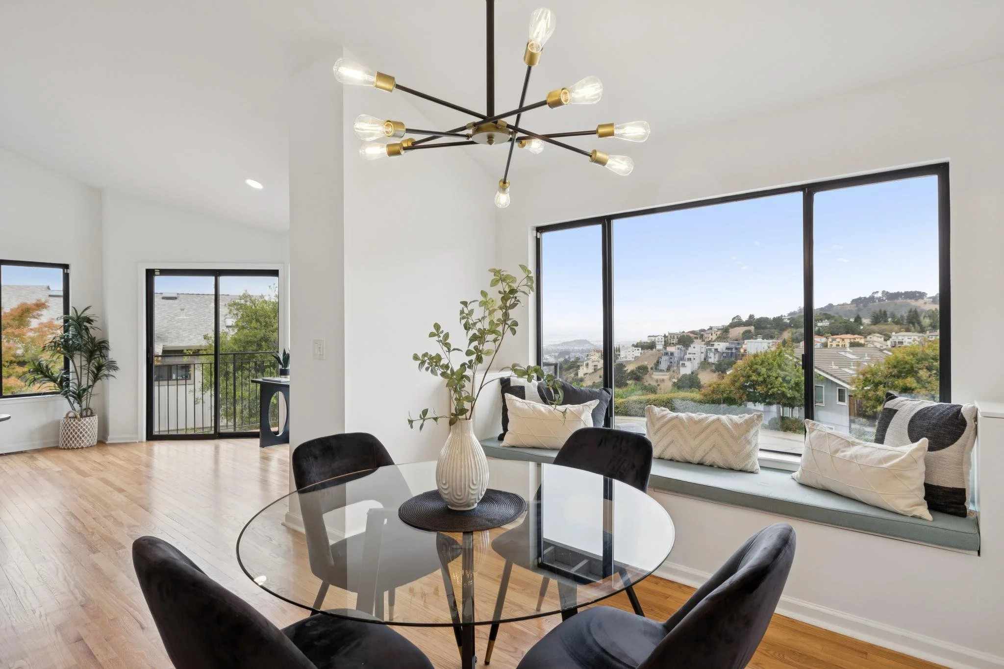 Modern living room with large windows showing a hillside neighborhood, a black chandelier with exposed bulbs, a round glass dining table with black chairs, and a white window seat with pillows and plants.