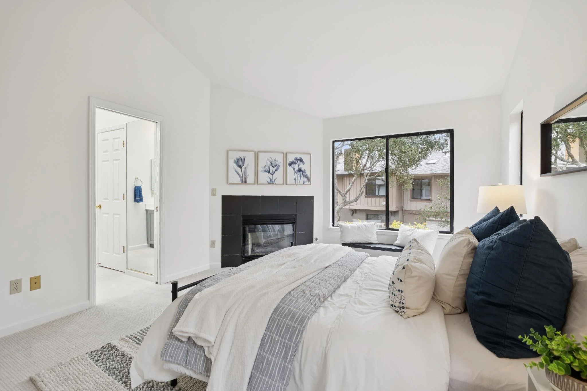 A bright bedroom with large windows, a bed with white and striped bedding, a dark blue pillow, framed floral art above a black fireplace, and a lamp on a side table.