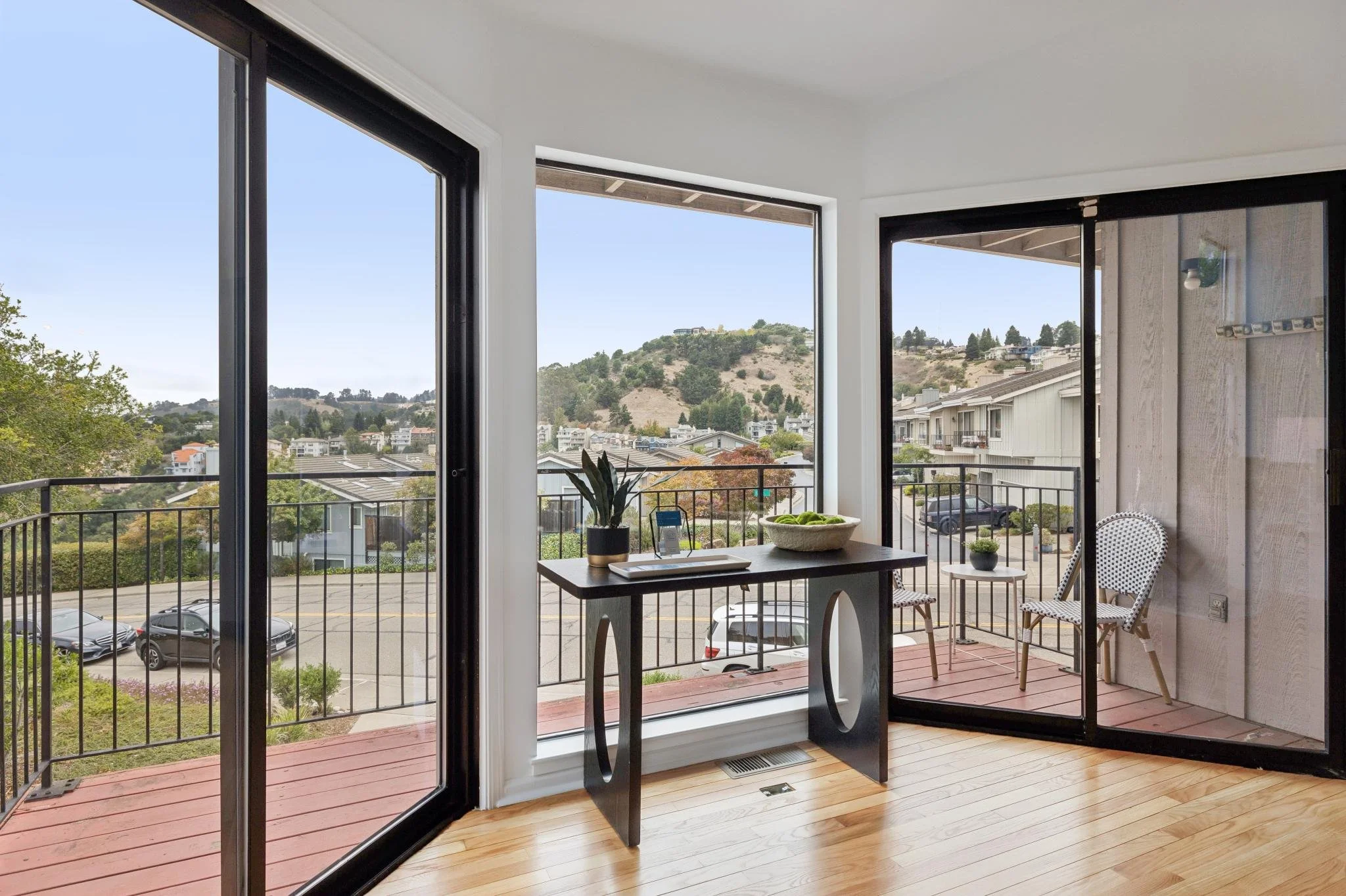 View of a balcony with a black railing and outdoor chairs, overlooking a parking lot, residential buildings, and a hillside with trees and houses.