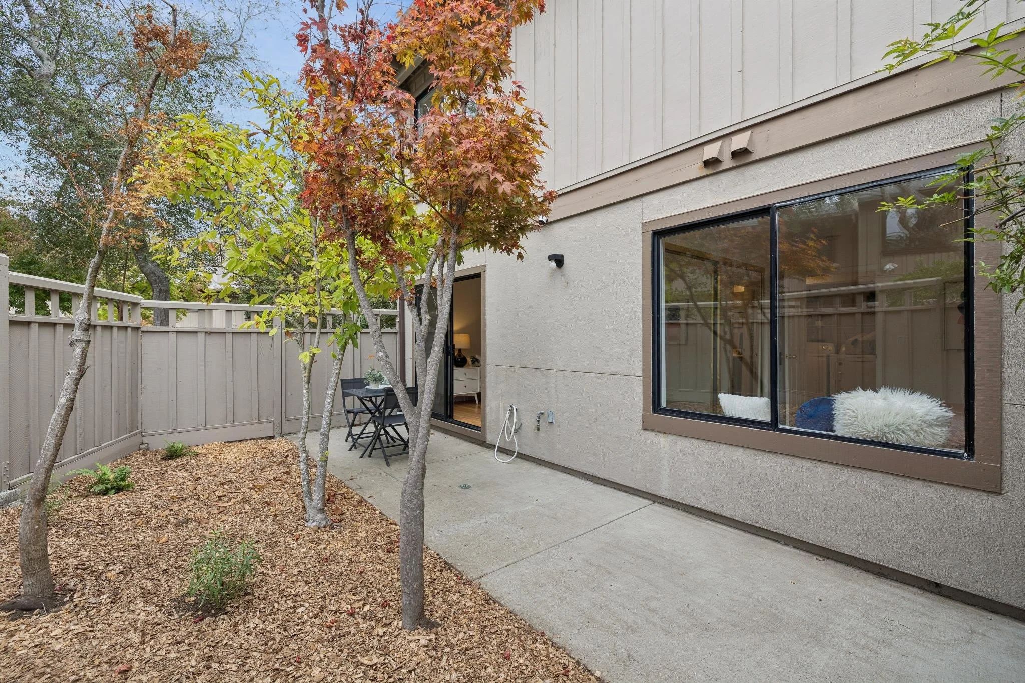Backyard patio area with trees, a beige house wall with large window, and outdoor furniture.