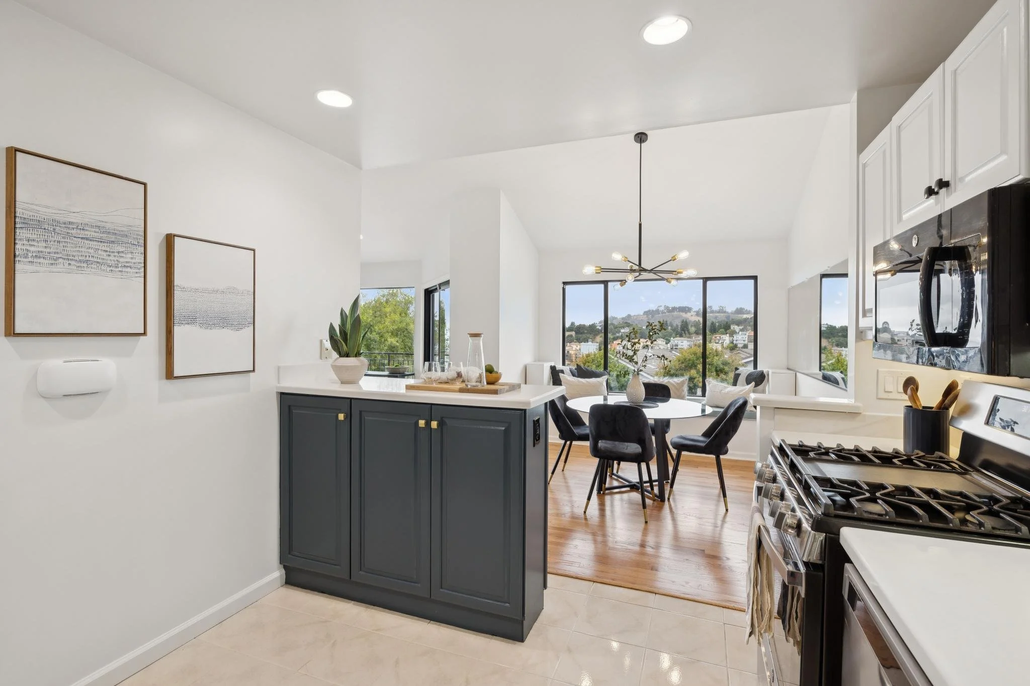 Interior of a modern kitchen and dining area with large windows showing a hillside view, featuring white walls, dark cabinets, a stainless steel stove, a black dining table with black chairs, and contemporary lighting fixture.