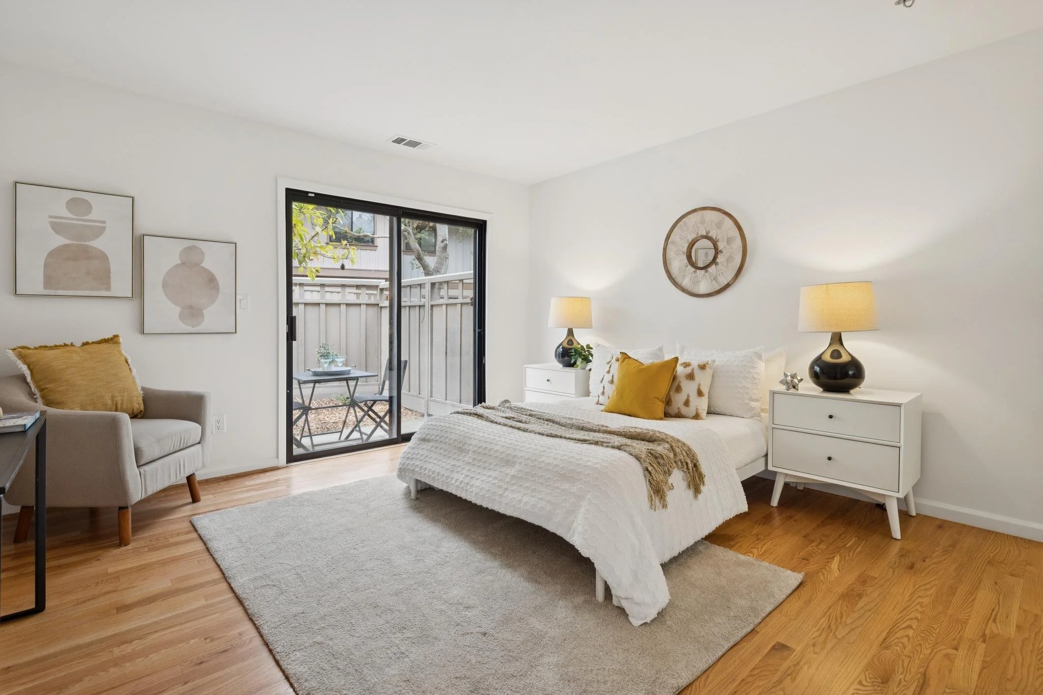 A bedroom with white walls, a bed with white bedding and pillows, two nightstands, matching lamps, and wall art, with a sliding glass door leading to an outdoor patio.