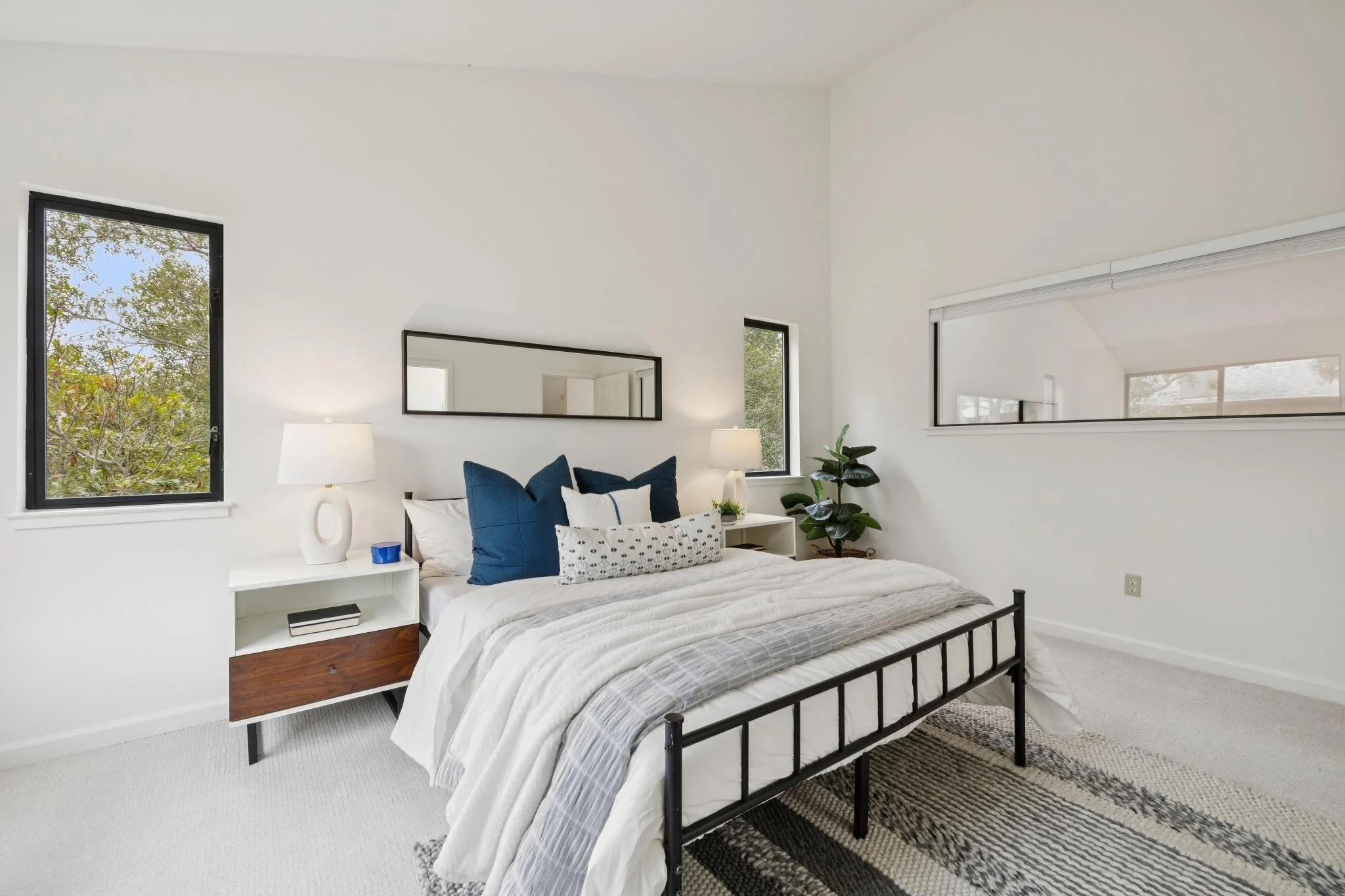 Modern bedroom with white walls, black metal bed frame, white and blue bedding, two windows, a large mirror, a white side table with a lamp, and a potted plant.