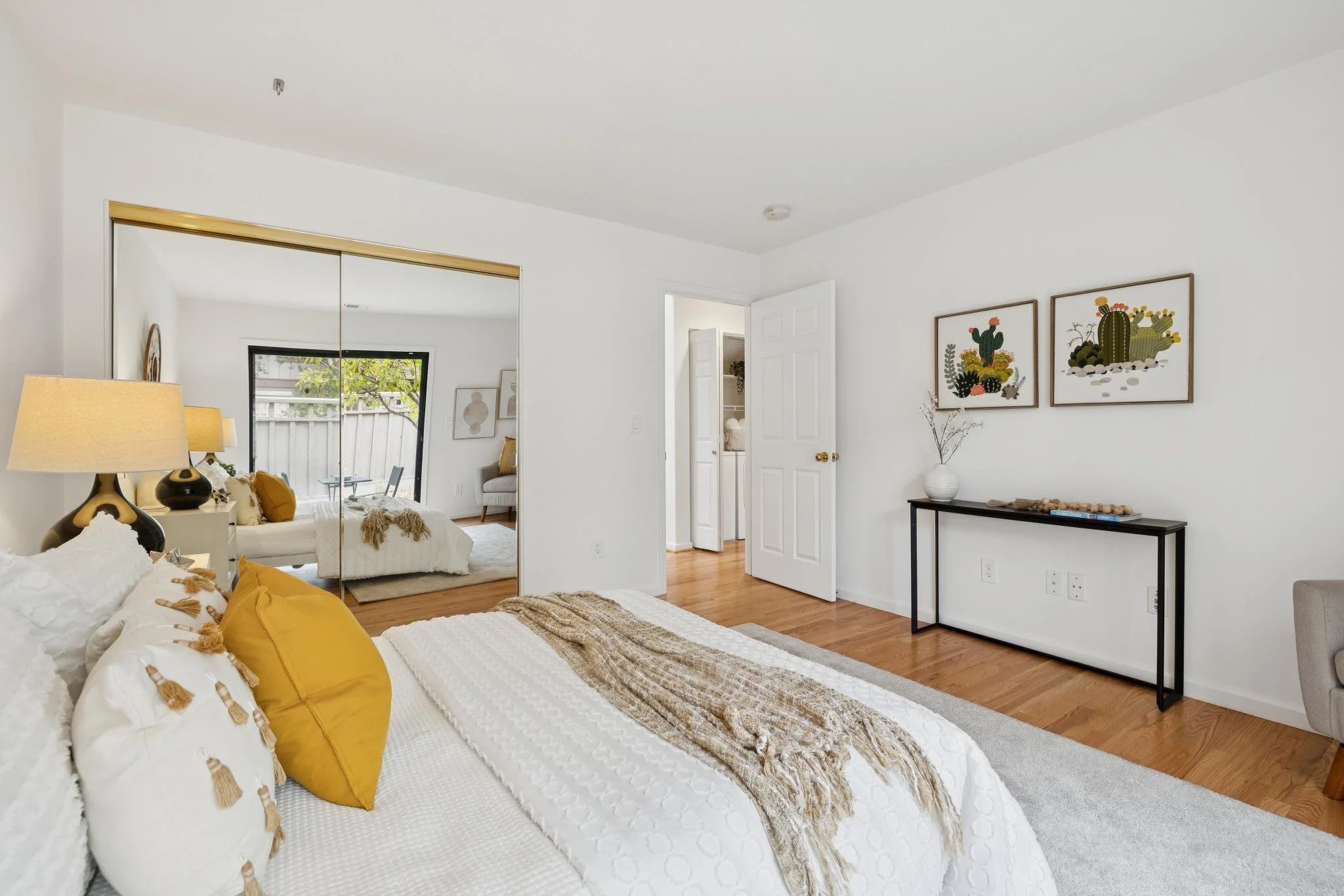 A bedroom with white walls, hardwood floors, a bed with white bedding and yellow pillows, and wall art with cacti designs.