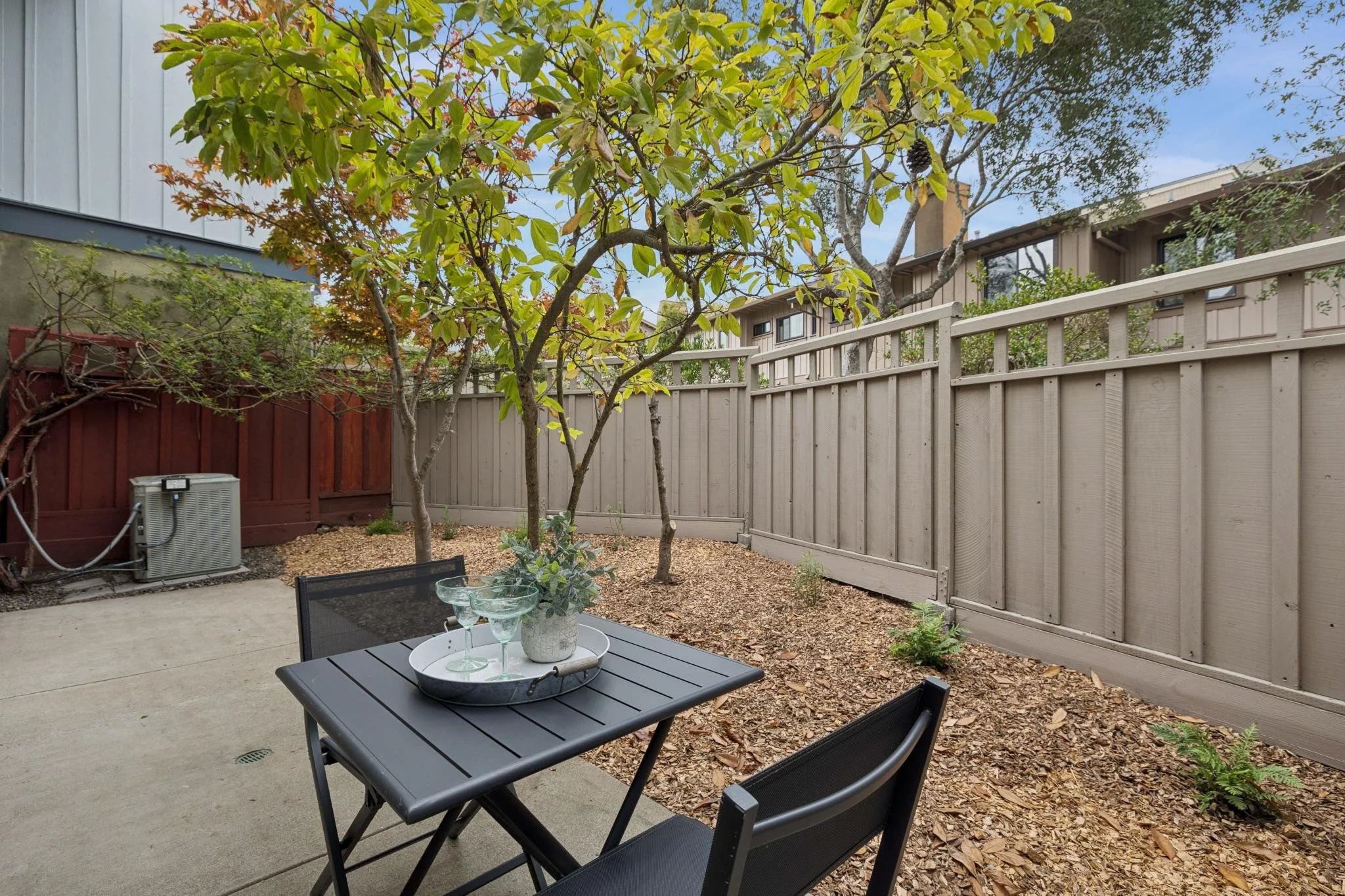 A backyard patio with black outdoor furniture including a table and chairs, a tray with glasses and a plant, surrounded by a garden with trees, some with autumn leaves, a beige wooden fence, and a two-story house in the background.