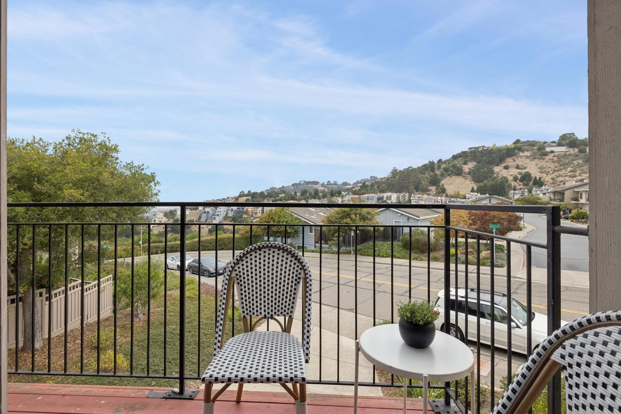 A balcony view overlooking a parking lot, trees, and a hillside with buildings and houses under a partly cloudy sky. There are two chairs and a small table with a potted plant on the balcony.