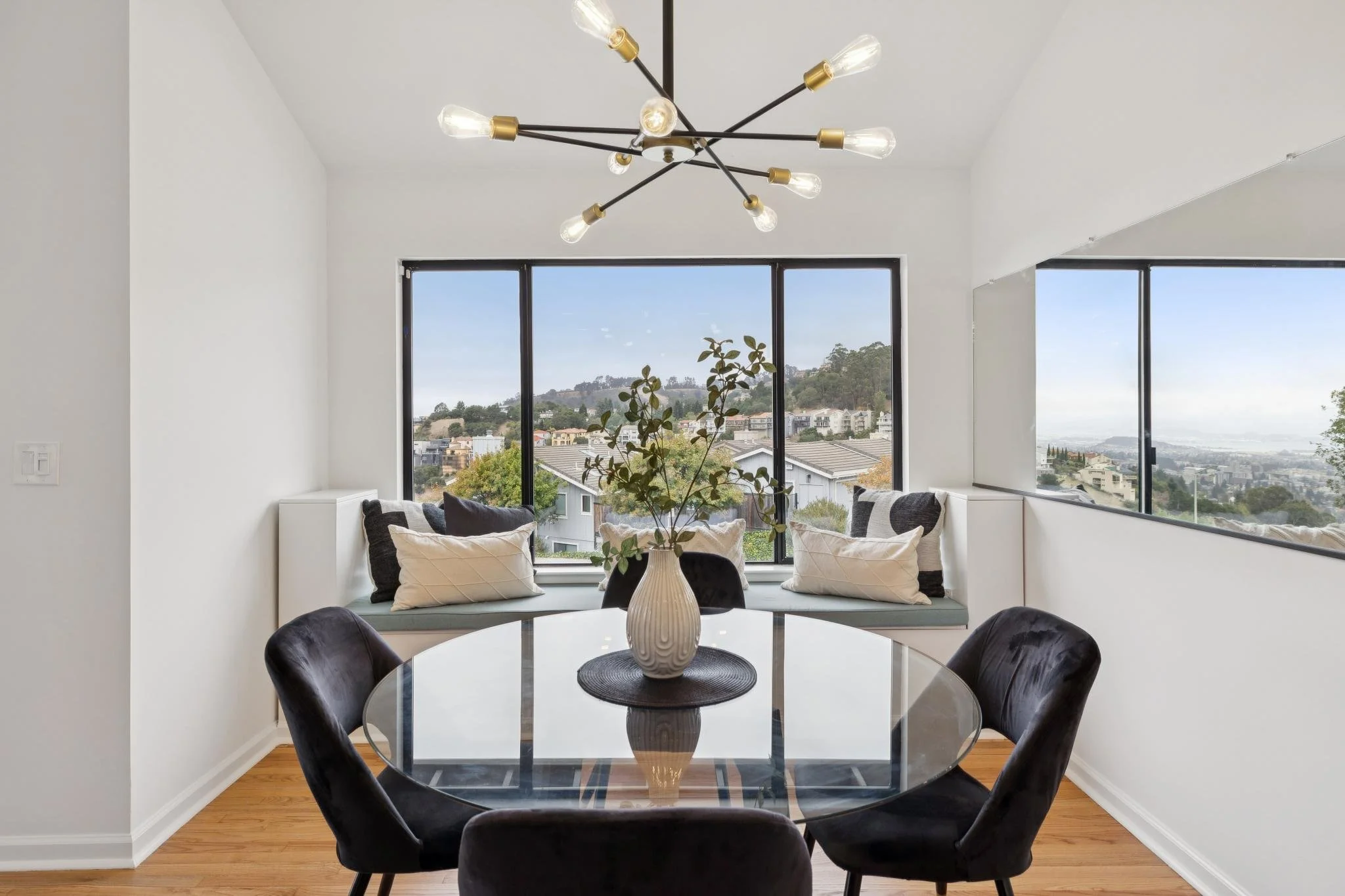 Contemporary dining room with round glass table, black velvet chairs, and a white vase with greenery centerpiece. Large windows reveal a hillside neighborhood view. Modern black chandelier with exposed lightbulbs.