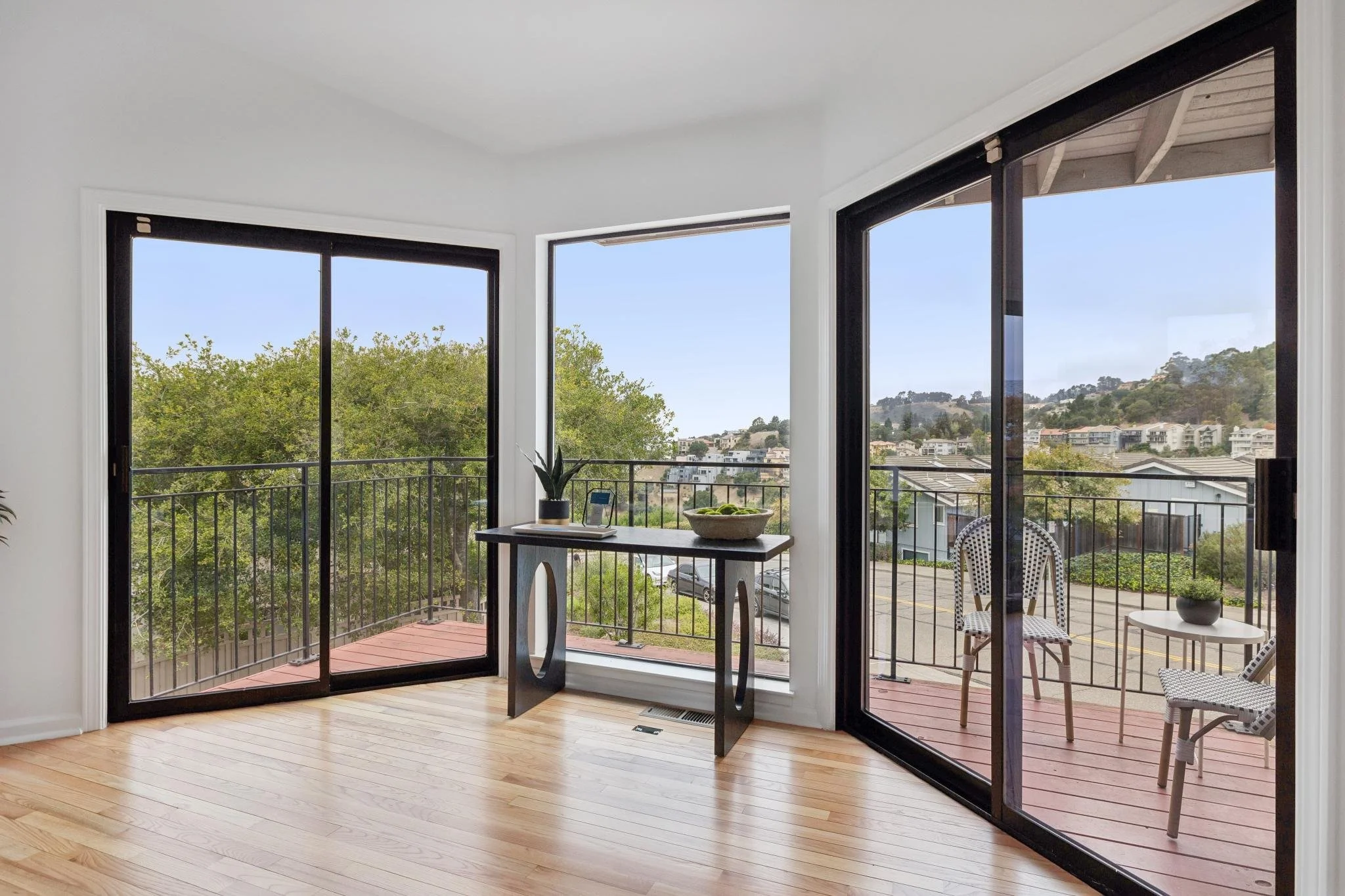 Bright sunlit living room with wooden floor and large glass sliding doors leading to a balcony, showing a view of trees and hillside homes outside.