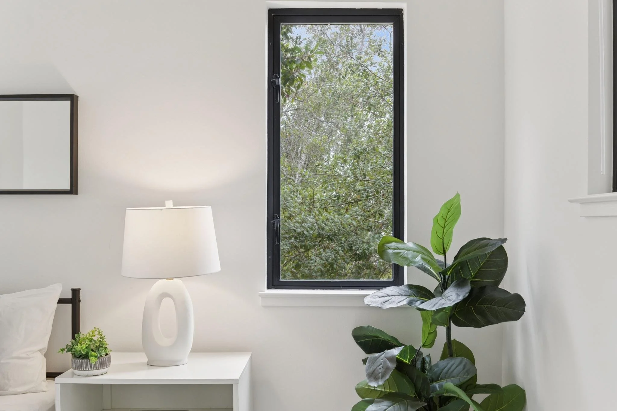Interior of a modern room with a black-framed window showing green trees outside. There is a white side table with a unique white ceramic vase and a small green potted plant, a white lamp, and a bed with white linens. Also, there is a green leafy pla