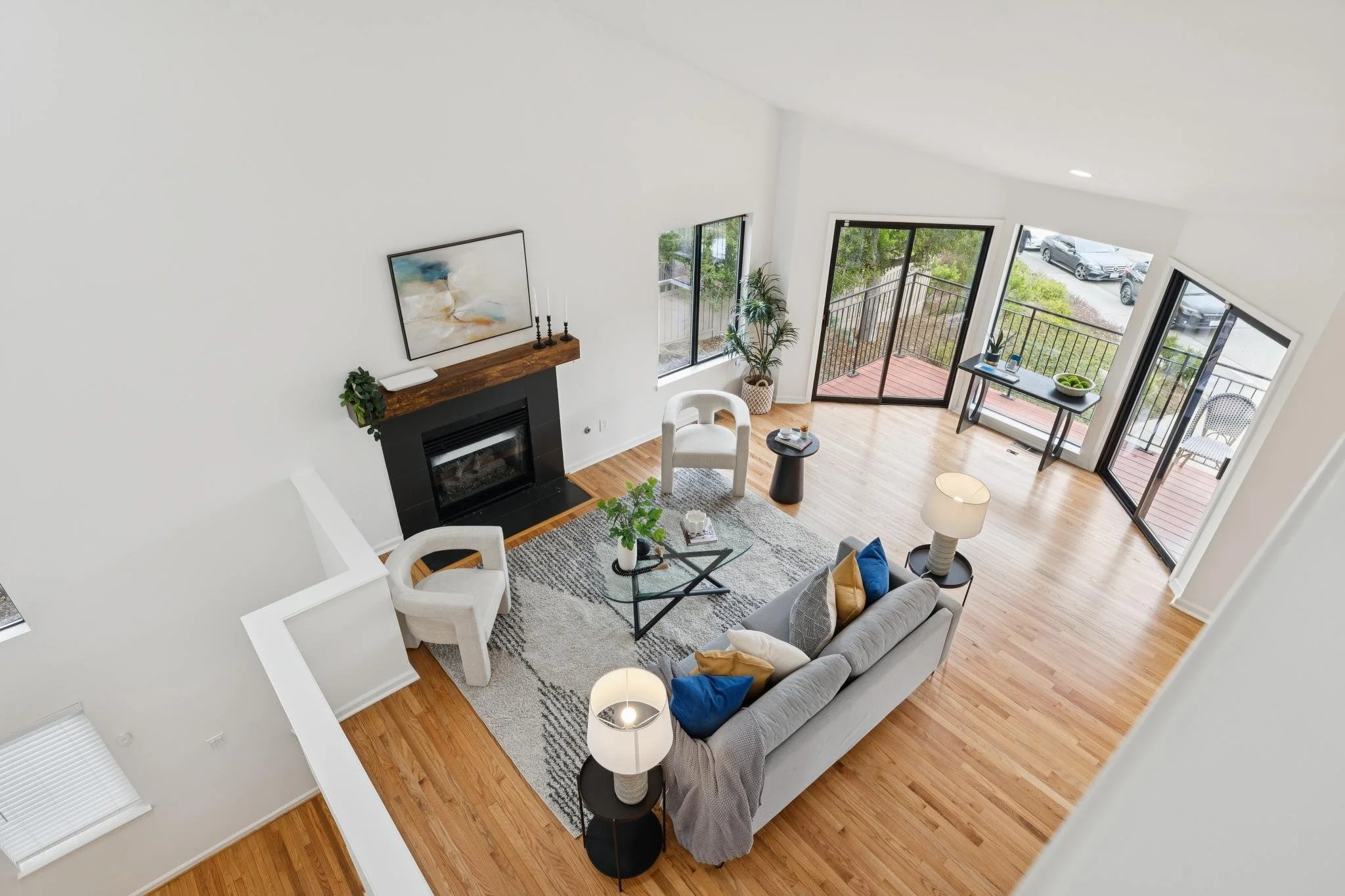 A spacious living room with white walls and hardwood flooring, featuring a gray sofa with colorful pillows, a white armchair, a black fireplace with a wooden mantle, paintings, and large windows with sliding glass doors leading to a balcony with outd