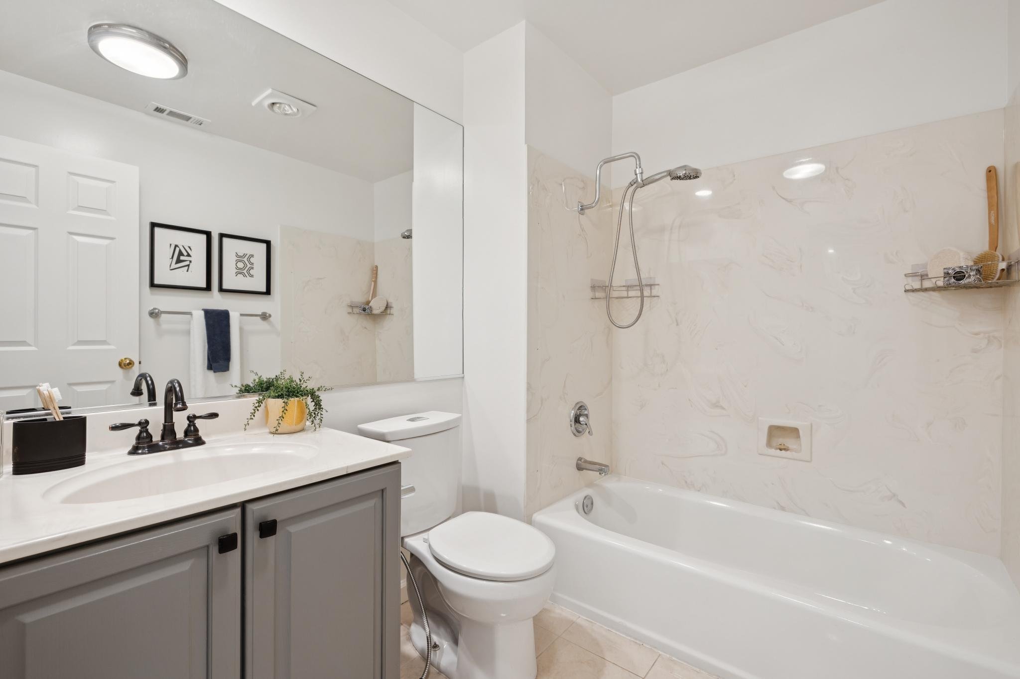 A clean bathroom with a white bathtub, a toilet, and a vanity with a sink and dark faucets. There are two framed art pieces on the wall, a towel rack with a dark towel, and a mirror above the sink. The bathroom has light-colored walls and a marble ac