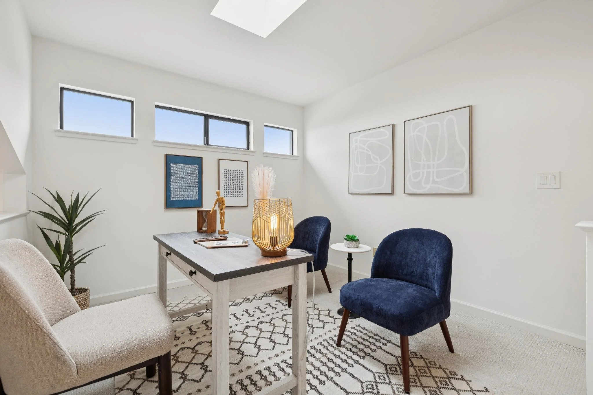 Bright living room features white walls, abstract art, a white rug with black geometric pattern, a beige armchair, dark blue velvet chairs, potted plants, and a table with decorative items and a lamp.