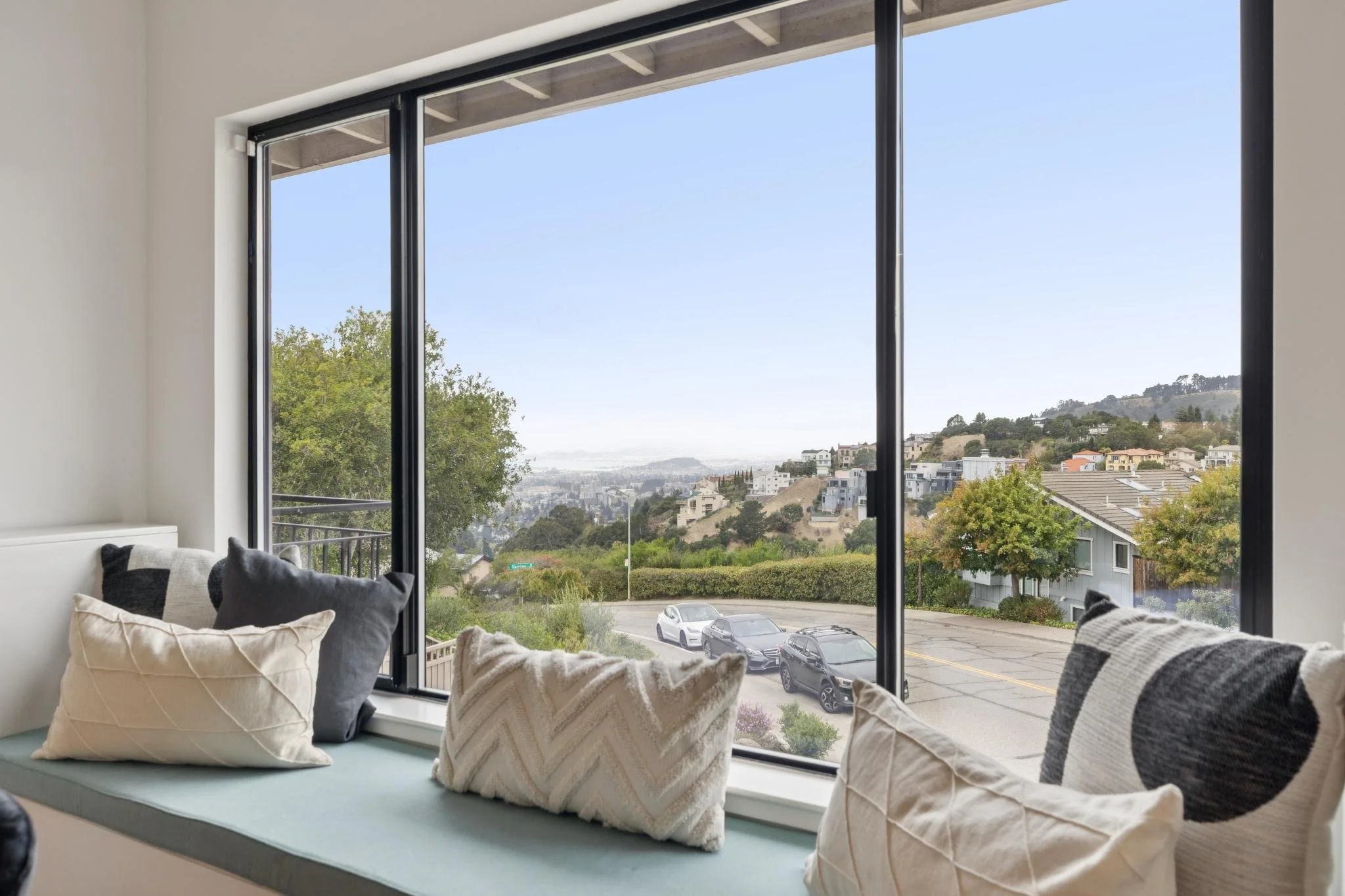 A window with a view of a hillside neighborhood with houses and parked cars, decorated with several cushions on the sill.