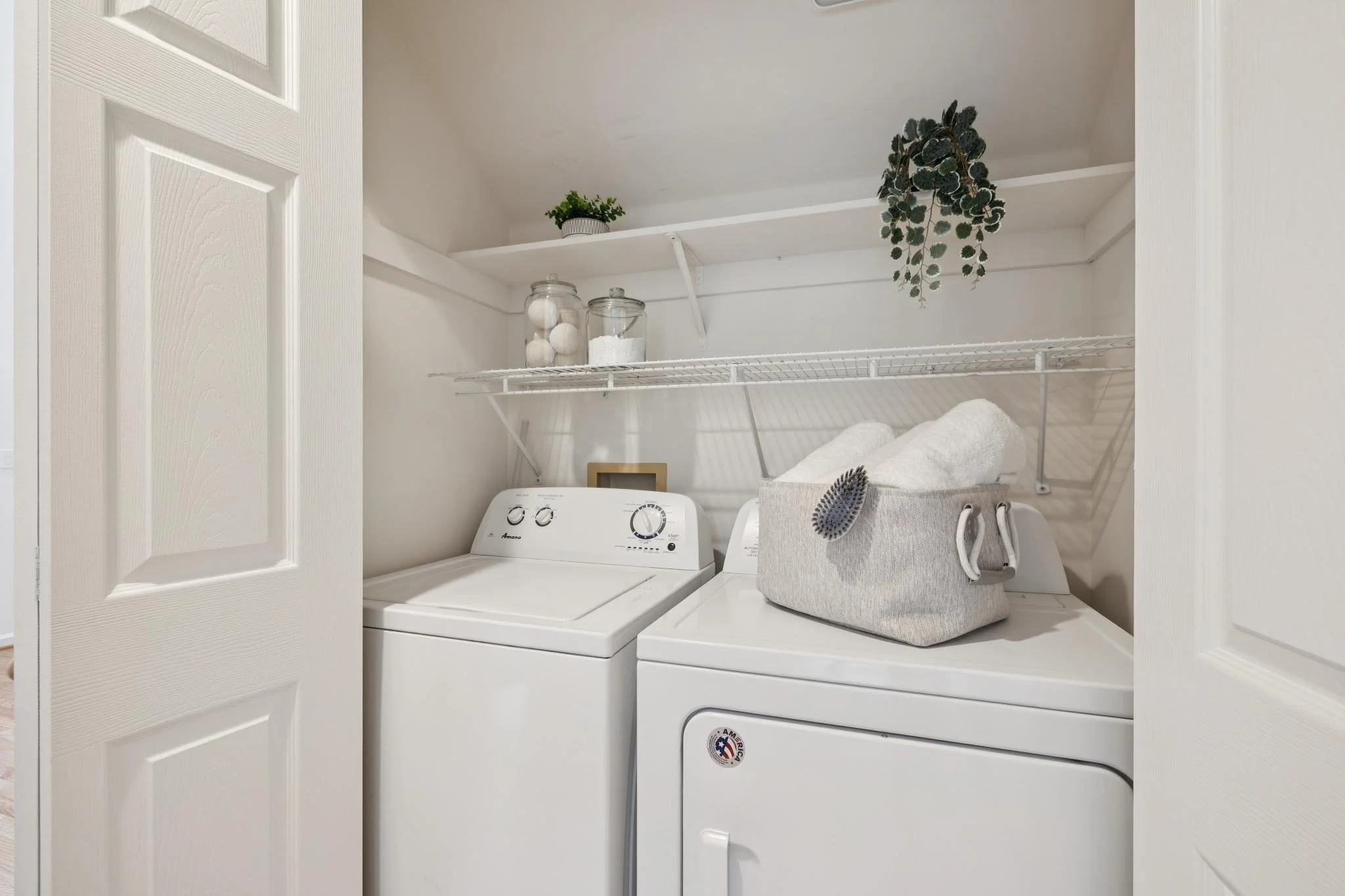 Inside a laundry closet with a top-loading washing machine and a front-loading dryer, containing shelves with decorative jars, a potted plant, and a hanging fern plant, surrounded by white walls and door.