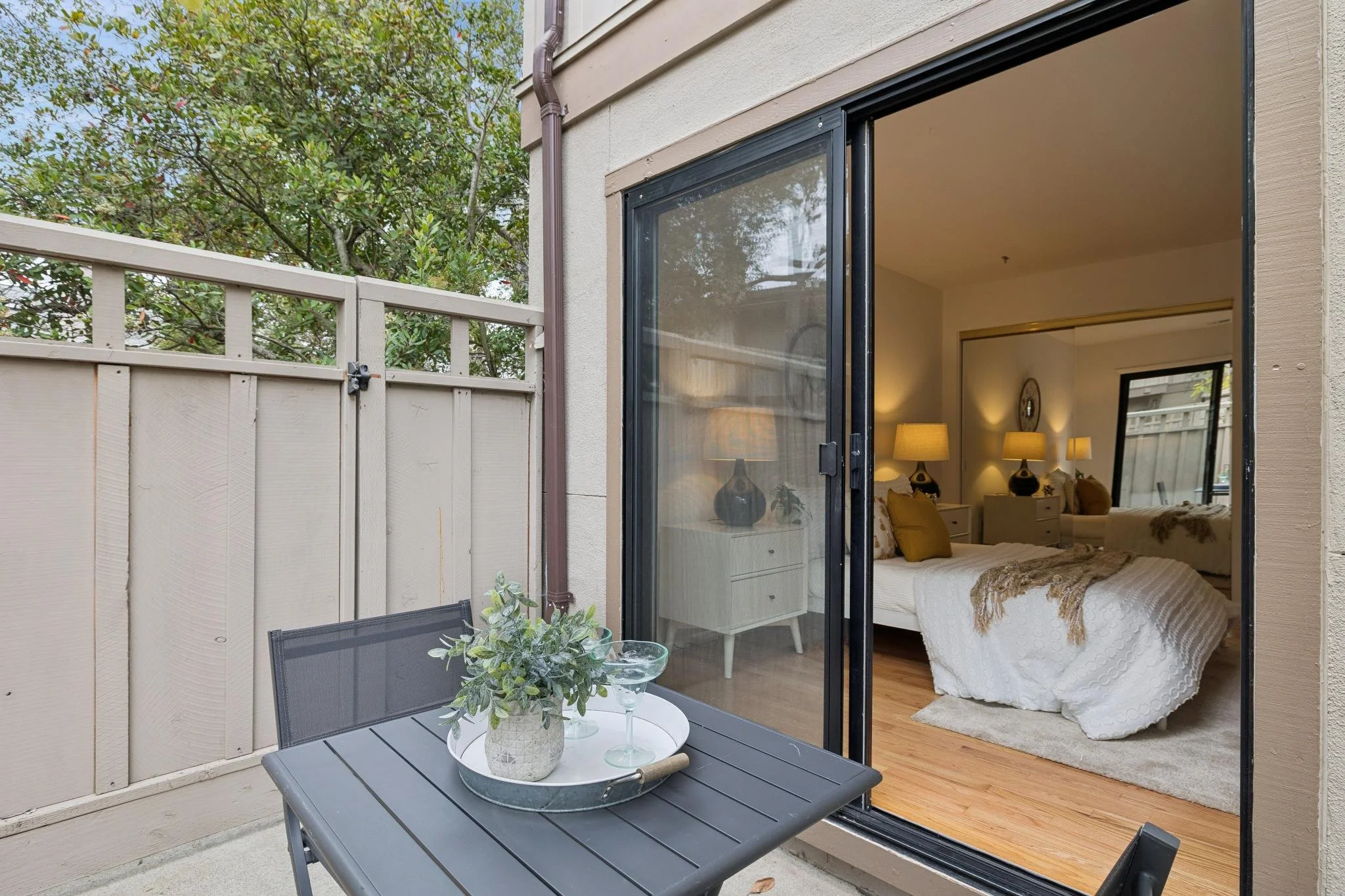 A view of a small outdoor patio area adjacent to a bedroom, seen through a sliding glass door. The patio has a gray table with a plant in a pot and a glass on it, and a black mesh chair. The bedroom interior shows a bed with a white quilt, pillows, a