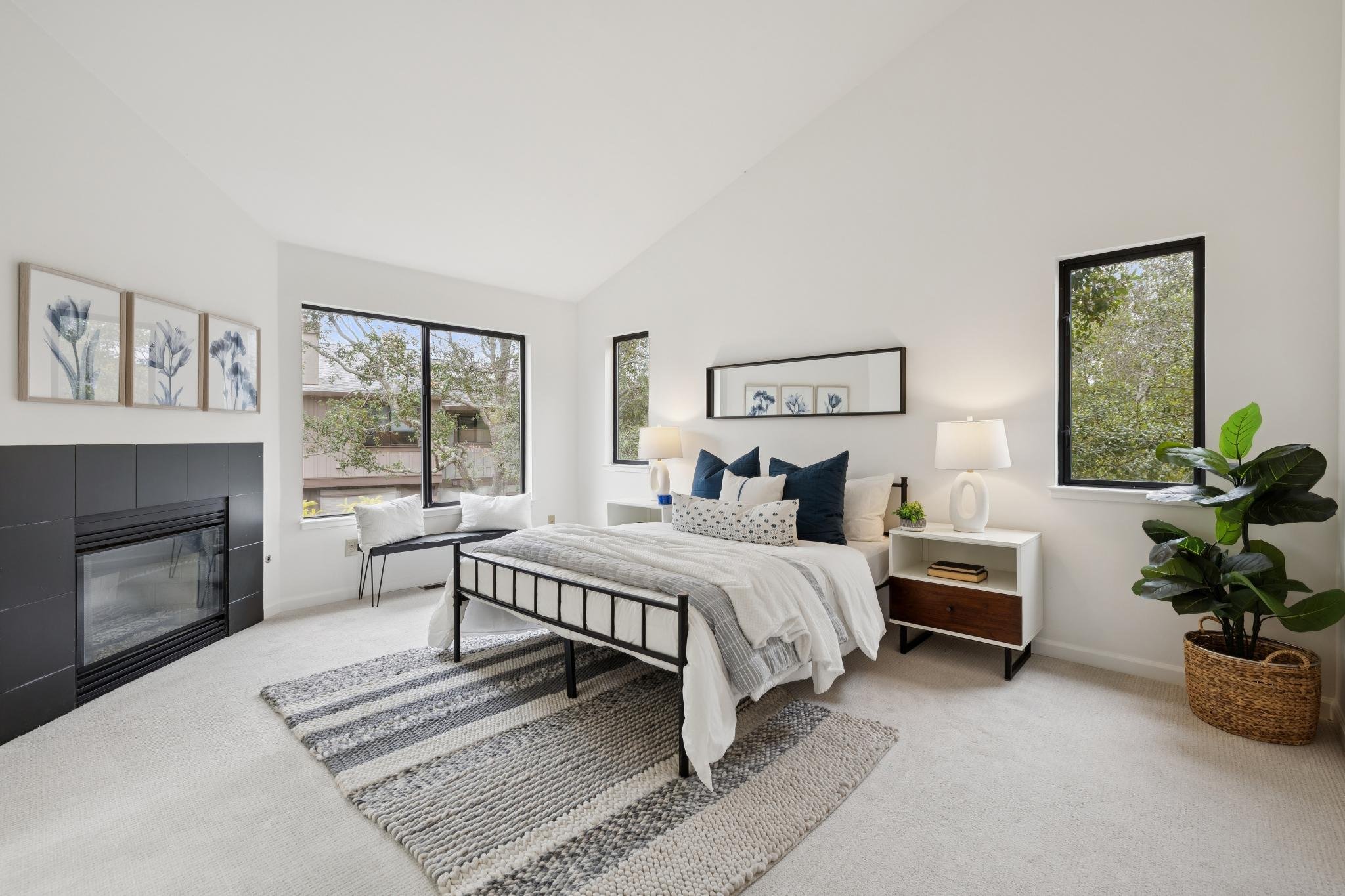 A bedroom with a white bed, black metal frame, and white bedding, flanked by matching nightstands with table lamps. There is a large potted plant in a woven basket in the corner. The walls are white with three framed blue flower artworks and a black 