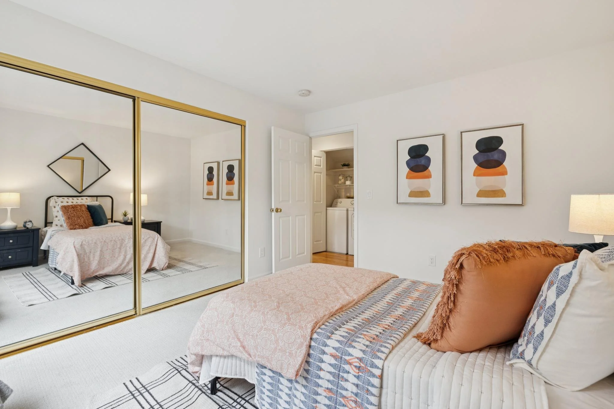 A bedroom with a bed, mirrored closet doors, framed artwork, and a view into a laundry room with a washer and shelves.