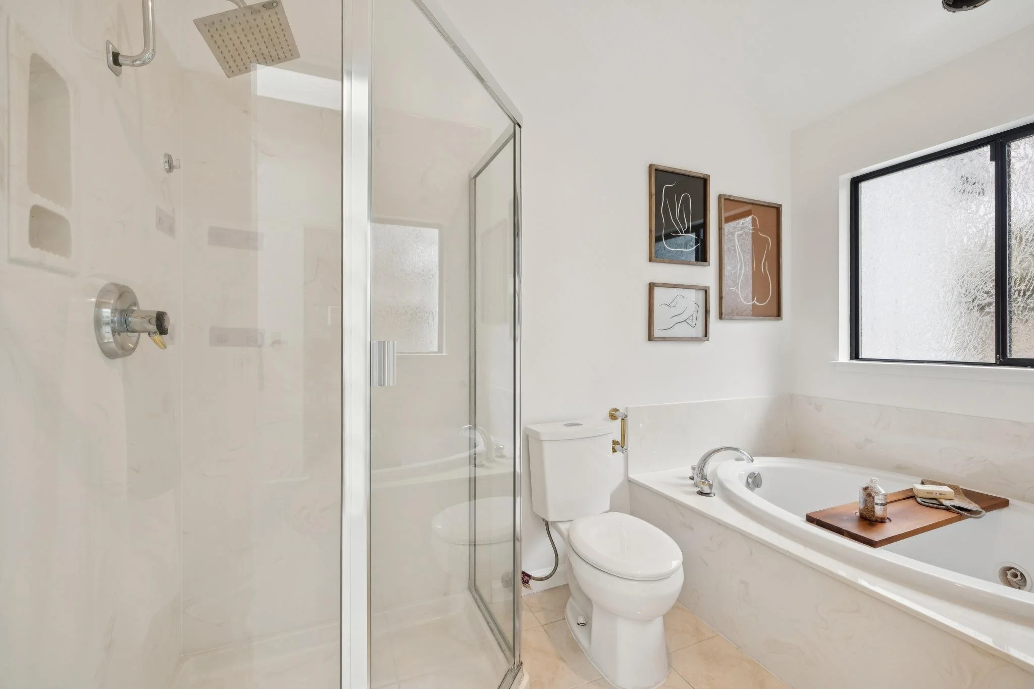A modern bathroom with a shower stall and a toilet. There are three framed line art pictures on the wall, a large frosted window, and a bathtub with a wooden tray holding toiletries.