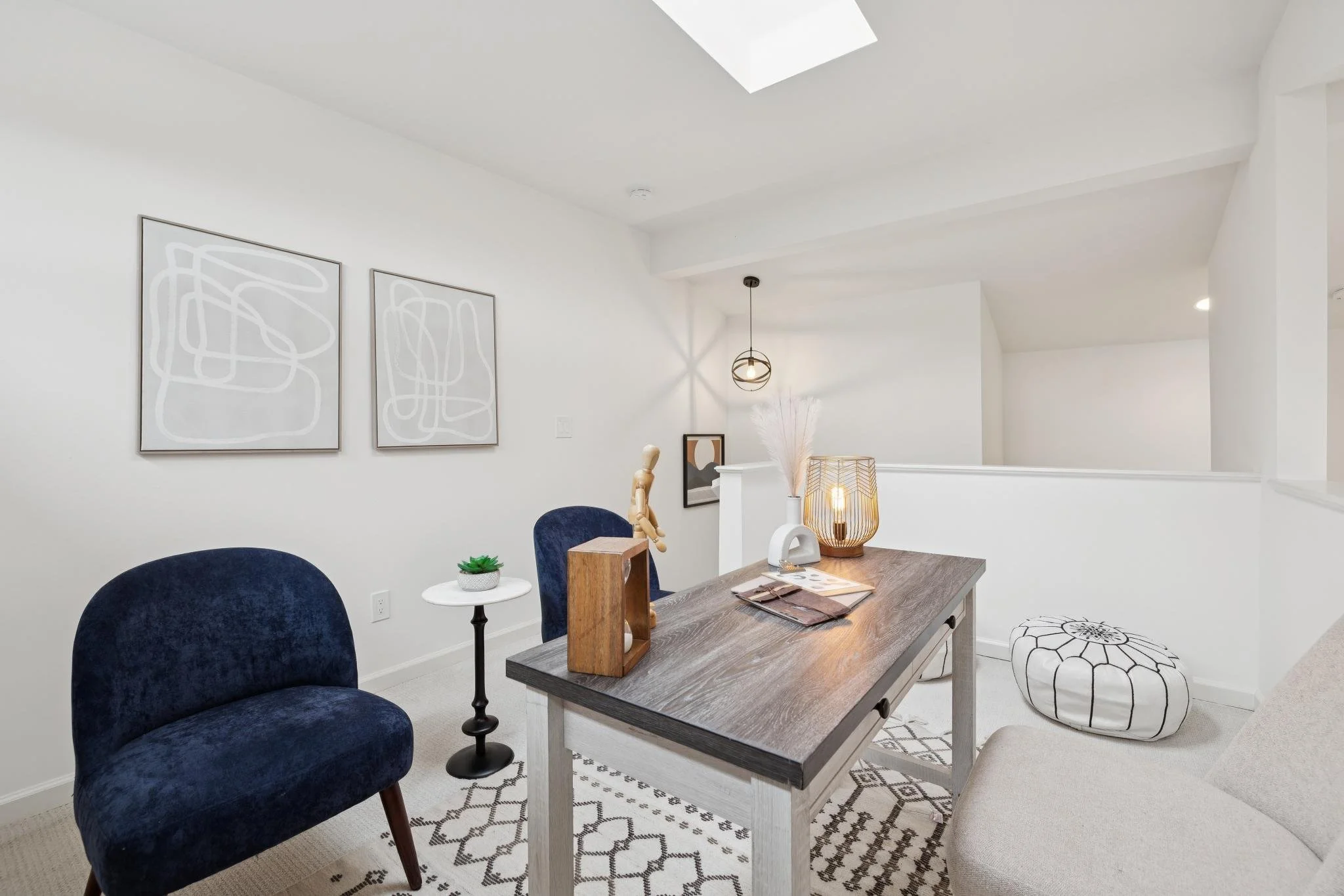 A bright, modern dining room with a wooden table, blue velvet chairs, white walls, and artwork. Decor includes a hanging light, a small white side table with a plant, and a table lamp. Skylight and patterned rugs complete the space.