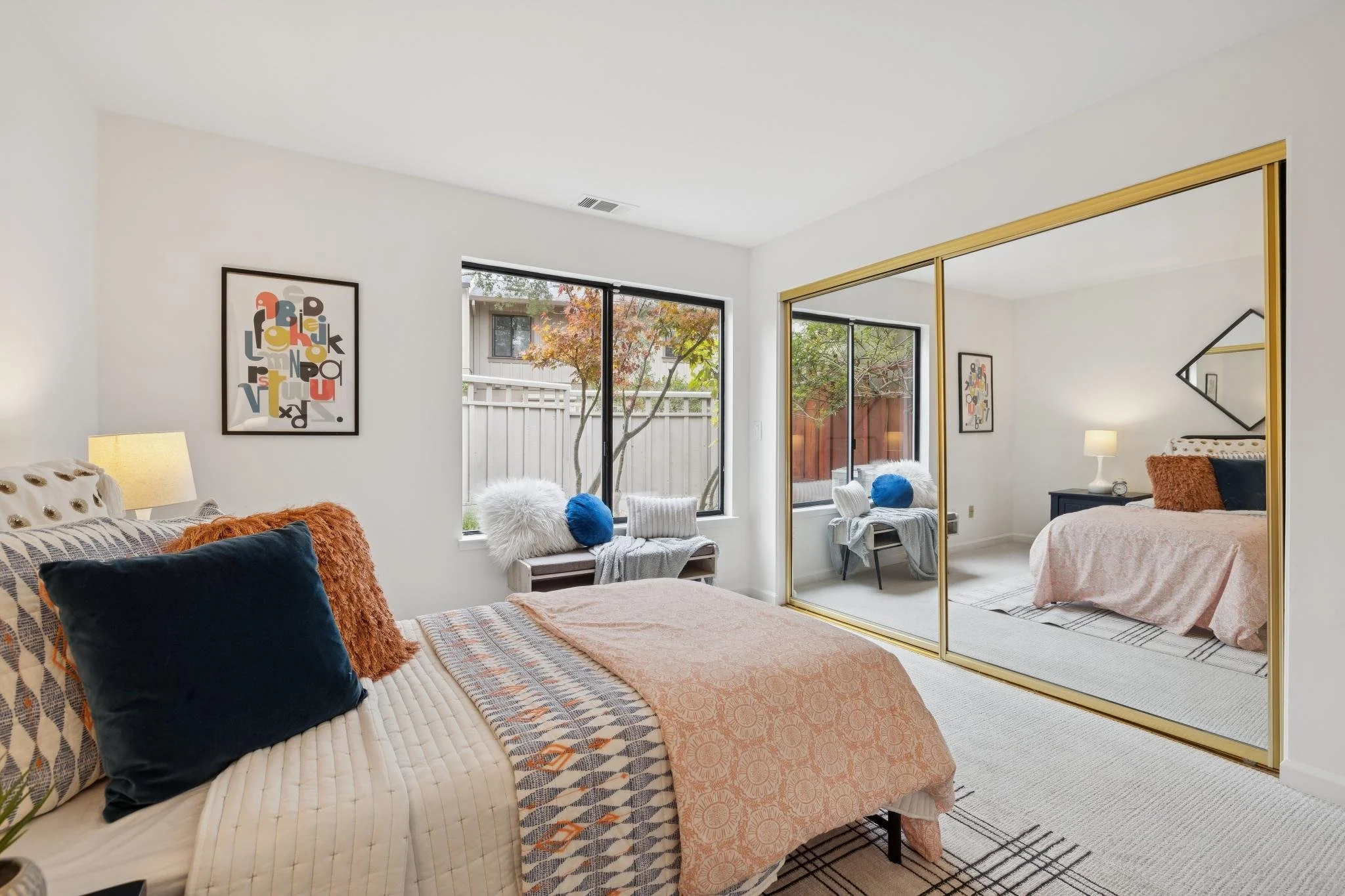 A bedroom with a bed covered in a beige quilt and colorful pillows, large windows with trees outside, a closet with mirrored sliding doors, and wall art.