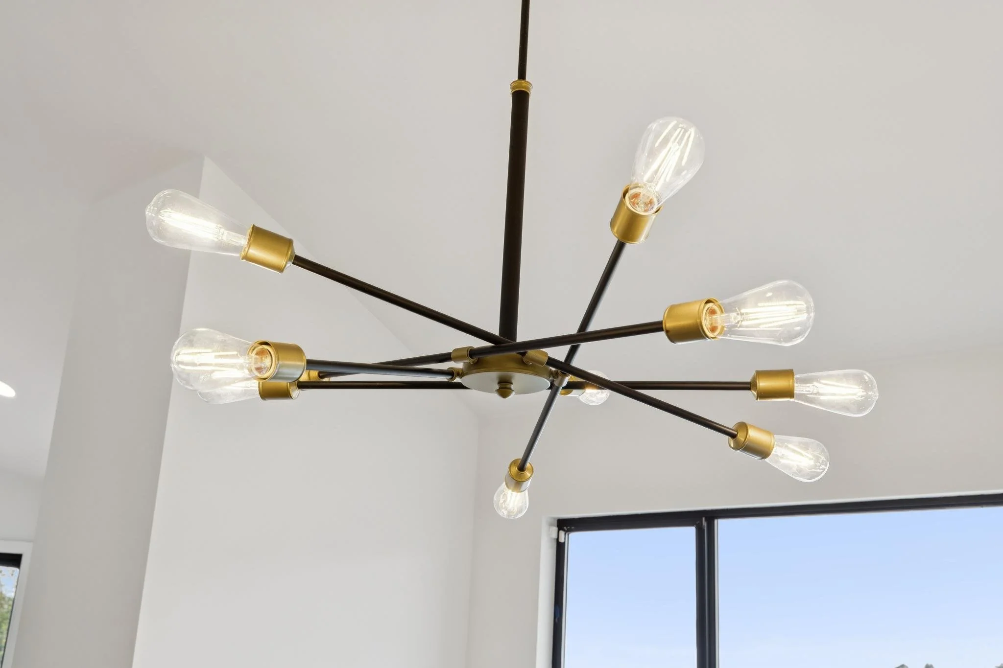 Modern chandelier with multiple exposed light bulbs hanging from black rods with gold accents, against a plain ceiling and a window showing a blue sky.
