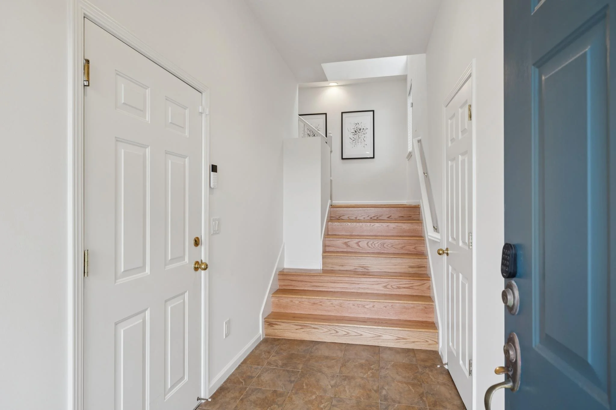 View of a house entryway featuring closed white double doors on the left, a blue front door on the right with a metallic handle, a keypad lock, and a doorbell. Straight ahead, a staircase with wooden steps and a white railing leads upward. Two framed