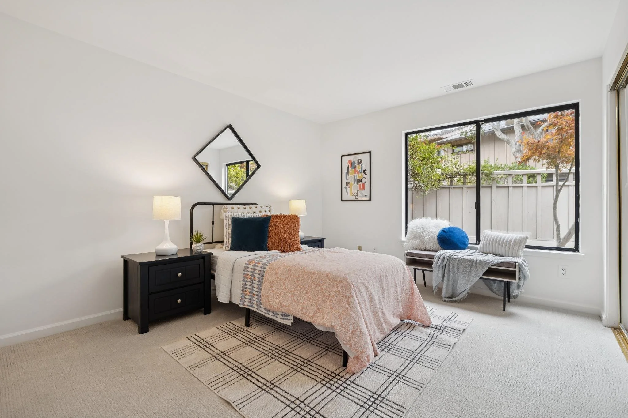 A bedroom with a bed, side tables, lamps, window, and artwork. The room has white walls, a beige carpet, and a patterned rug.