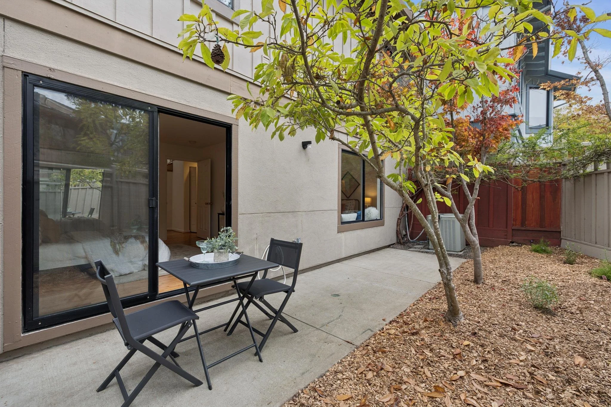 A backyard patio with a small round table set with drinks and plants, trees with autumn leaves, and large sliding glass doors leading into a house.