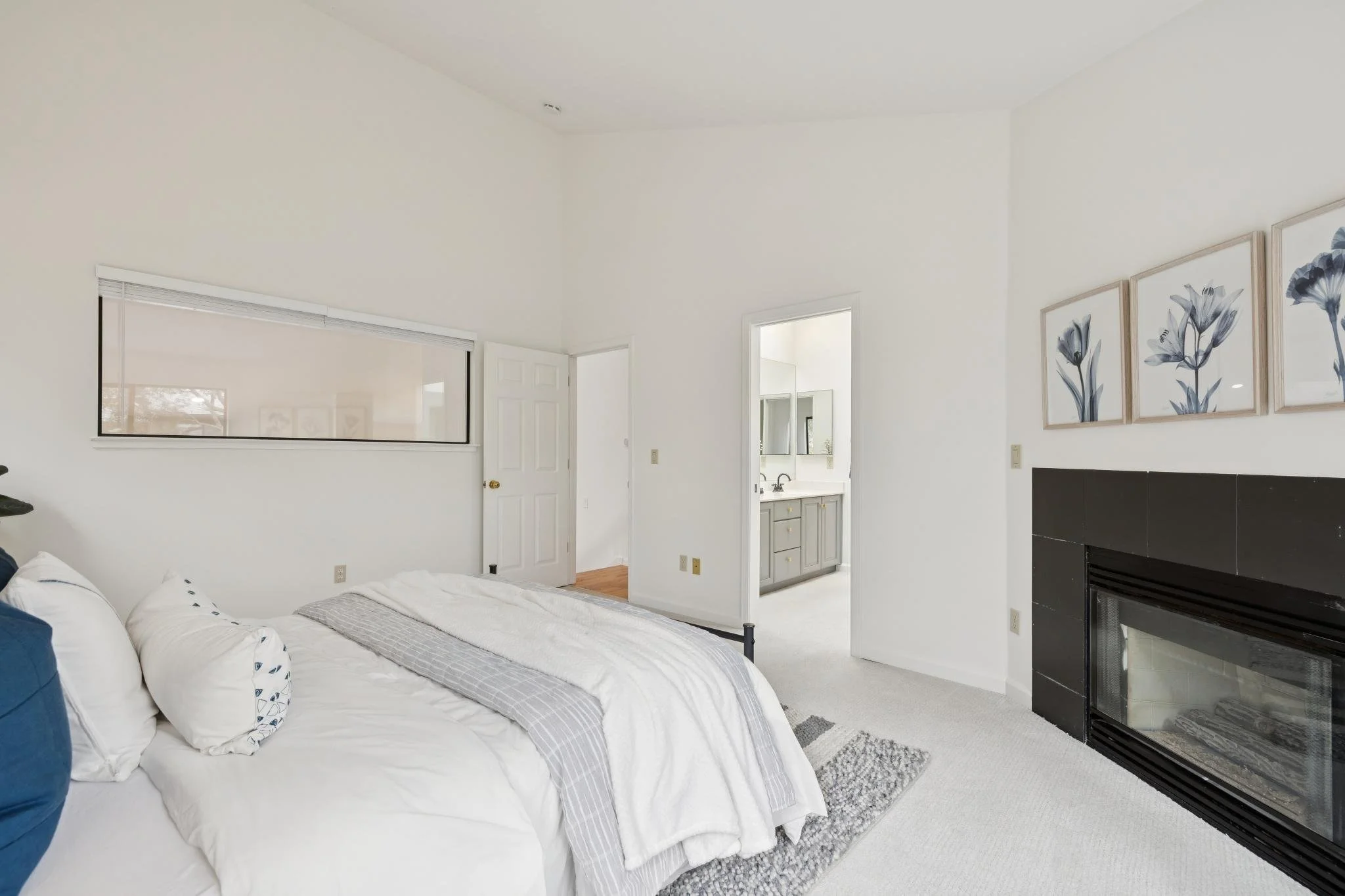 A bedroom with a white bed, decorative pillows, a gray blanket, and a rug on light-colored carpet. There is a black fireplace on the right and framed flower art on the white wall. A doorway shows a bathroom with gray cabinetry and a mirror.