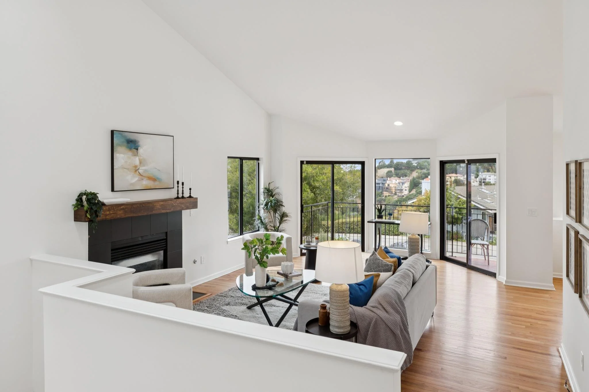 Bright living room with white walls, wooden floors, large windows, and seating area including a gray sofa, armchair, and coffee table, with view of cityscape outside.