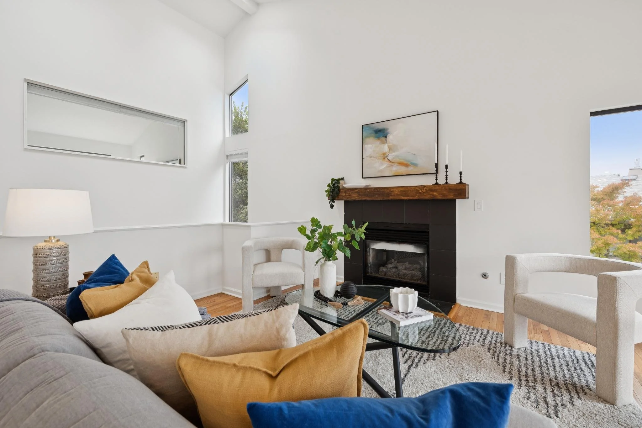 A modern living room with white walls, a fireplace with a wooden mantel, and abstract artwork hanging above. There are two white chairs, a sofa with colorful pillows, a glass coffee table with decorative items, and tall windows letting in natural lig