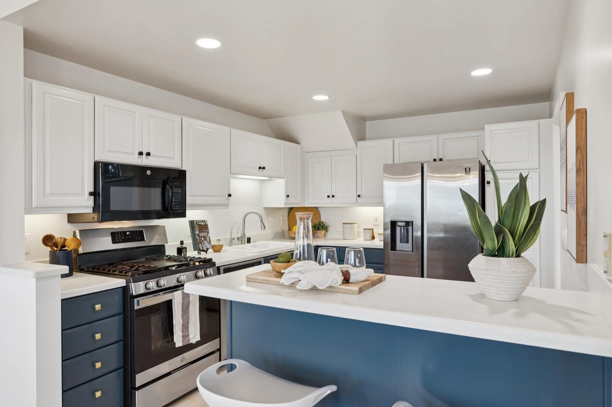 A modern kitchen with white upper cabinets, stainless steel appliances including a microwave and refrigerator, and a blue lower cabinet. There is a white kitchen island with a wooden cutting board, a white bowl with green fruit, a white cloth, and a 