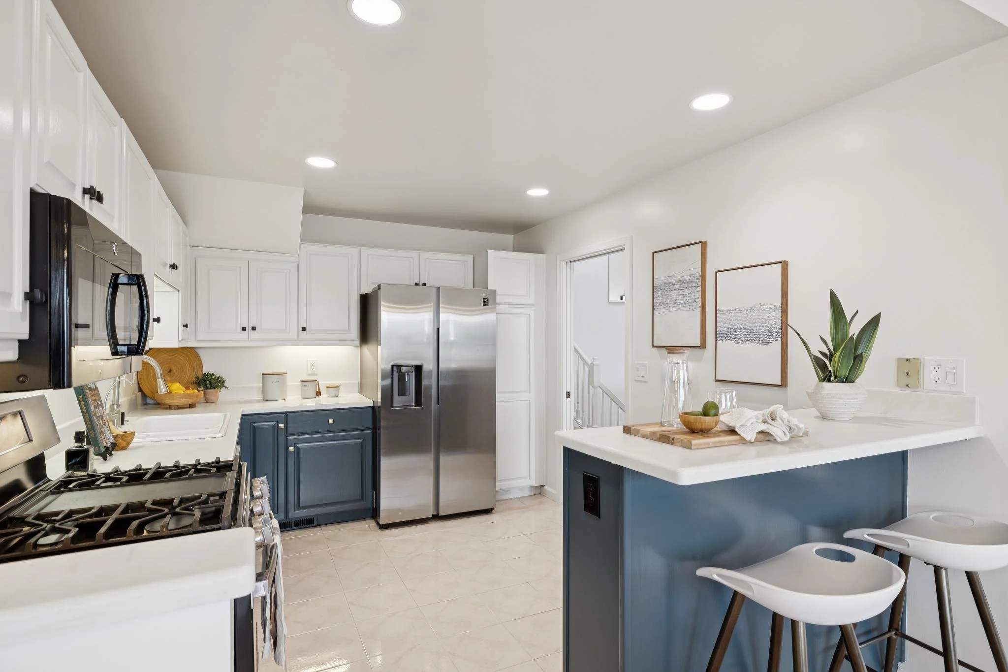 A modern kitchen with white upper cabinets, navy lower cabinets, stainless steel refrigerator, and a white countertop island with bar stools. Decor includes framed artwork, a potted plant, and kitchen utensils.