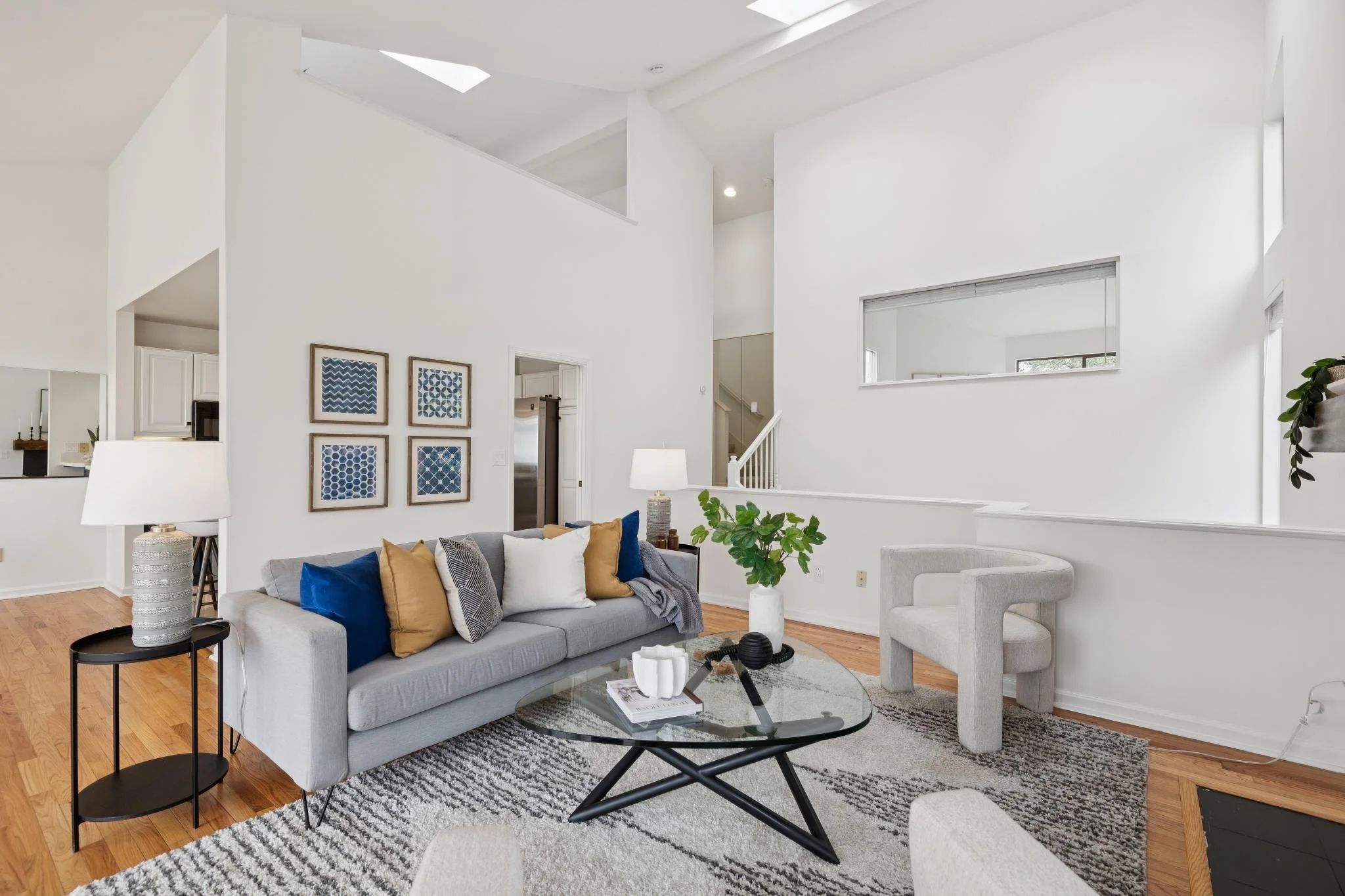A modern living room with a light gray couch adorned with various colored pillows, a glass coffee table, and a black side table with a white decorative vase. The room features white walls, a patterned black and white rug, and wooden flooring. There a