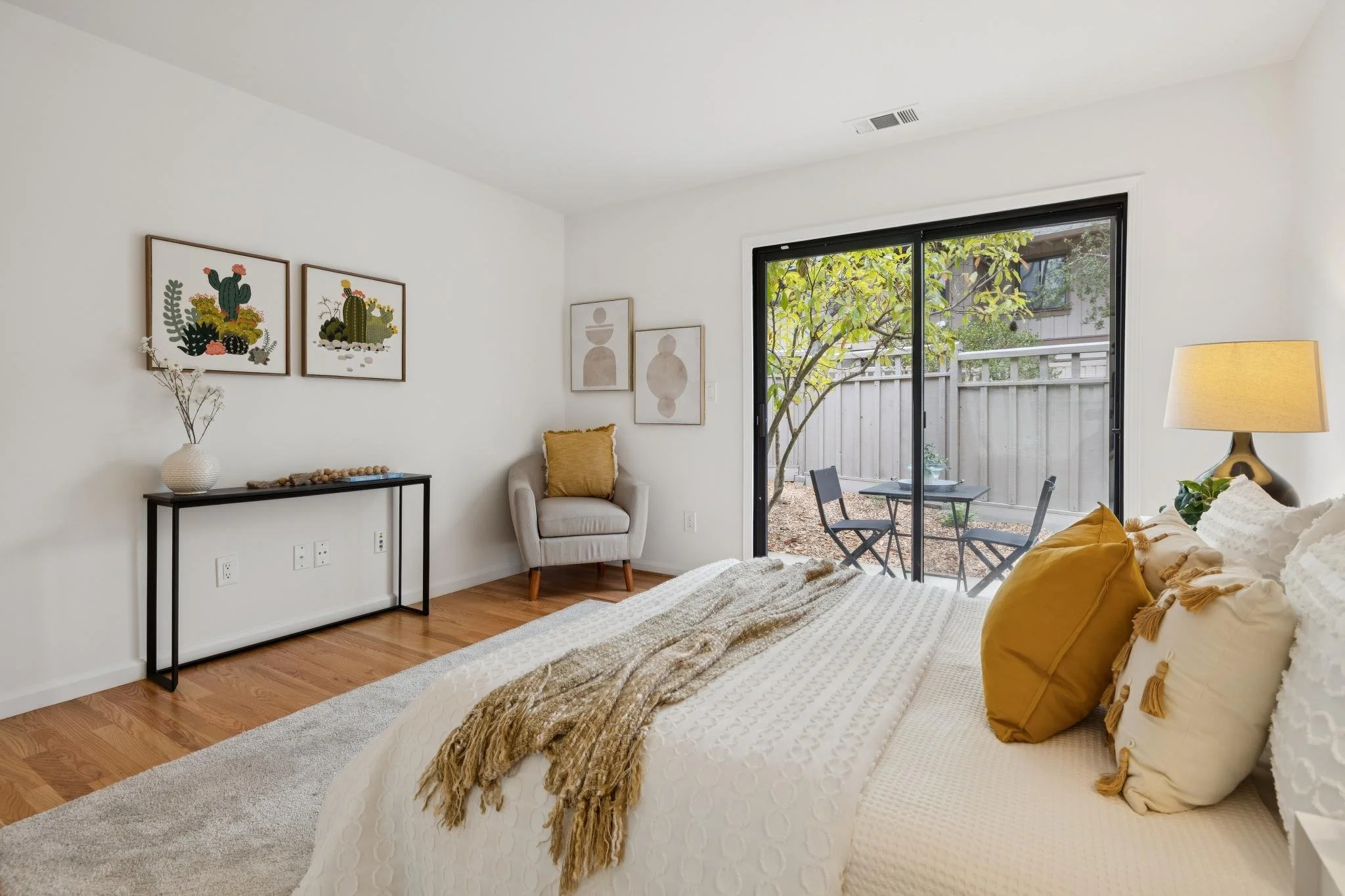 A cozy bedroom with a white bed, yellow pillows, and a textured blanket. A side table with a lamp, a beige armchair with a yellow pillow, and wall art featuring desert and abstract designs. A sliding glass door leads to an outdoor patio with a tree a