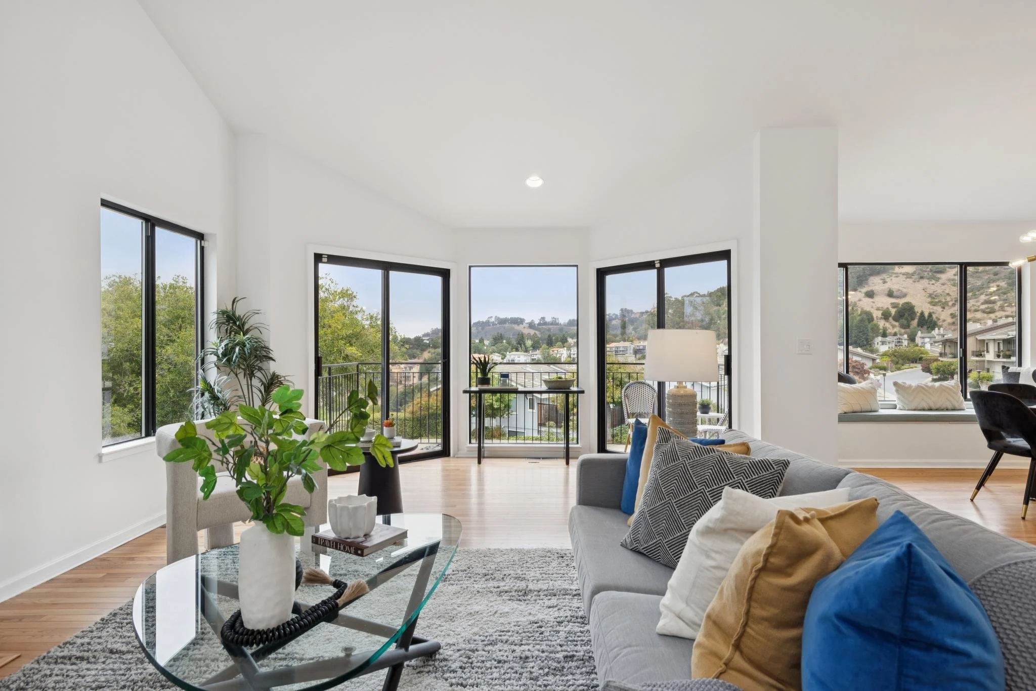 Bright living room with large windows showing a scenic view of hills and houses, modern furniture including a gray couch with patterned and solid pillows, a glass coffee table with decorative vases and books, and patterned armchairs, with hardwood fl