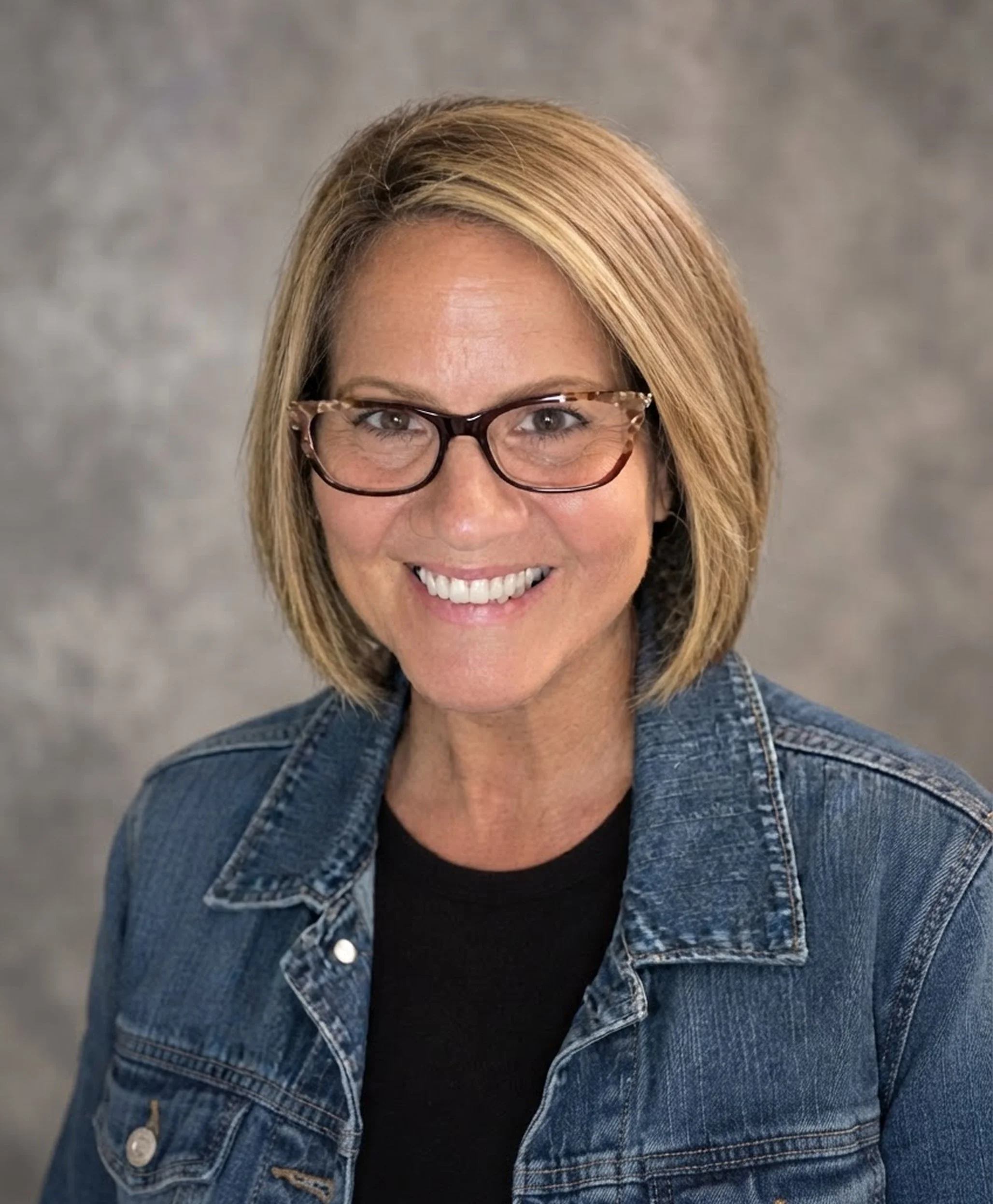 Close-up of a smiling woman with blonde bob haircut, wearing tortoiseshell glasses, a denim jacket, and a black top, against a grey background.