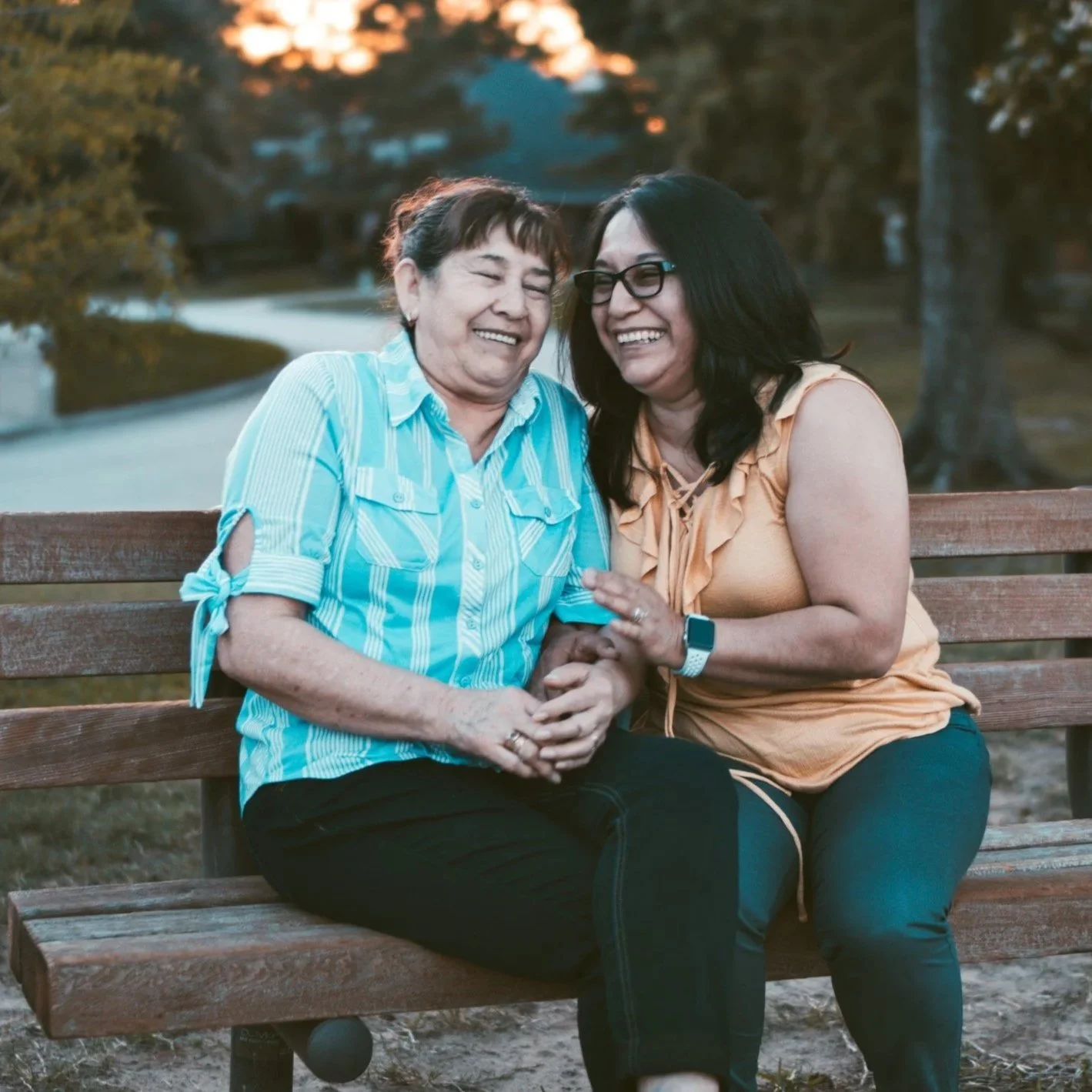 Two women sit closely on a park bench, smiling and laughter shared between them, during sunset with trees and a walking path in the background.