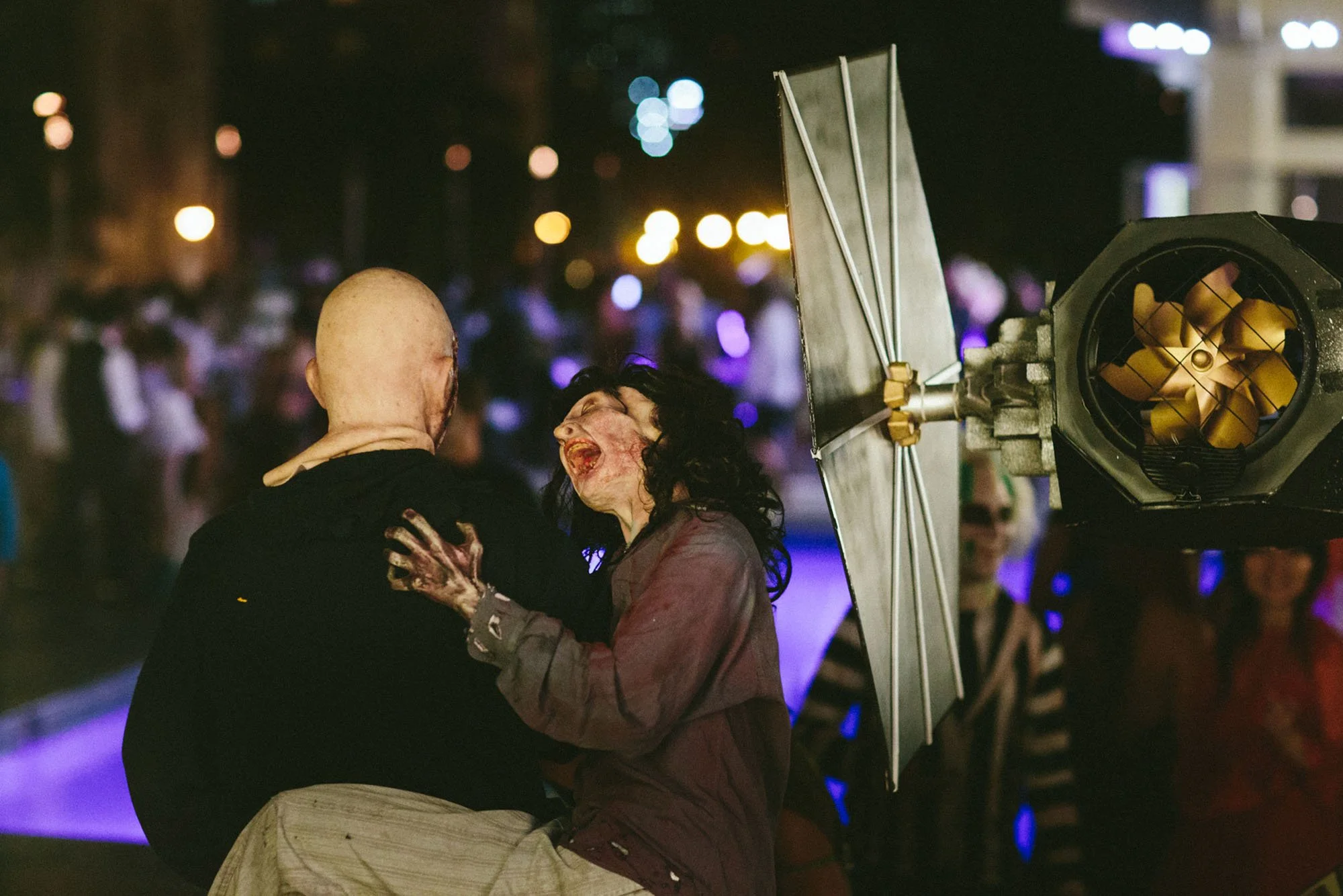 A person with zombie makeup and costume holds the face of another person, who is wearing a black jacket, in front of a large film or stage light at night. The scene is dimly lit with colorful, out-of-focus city lights in the background.