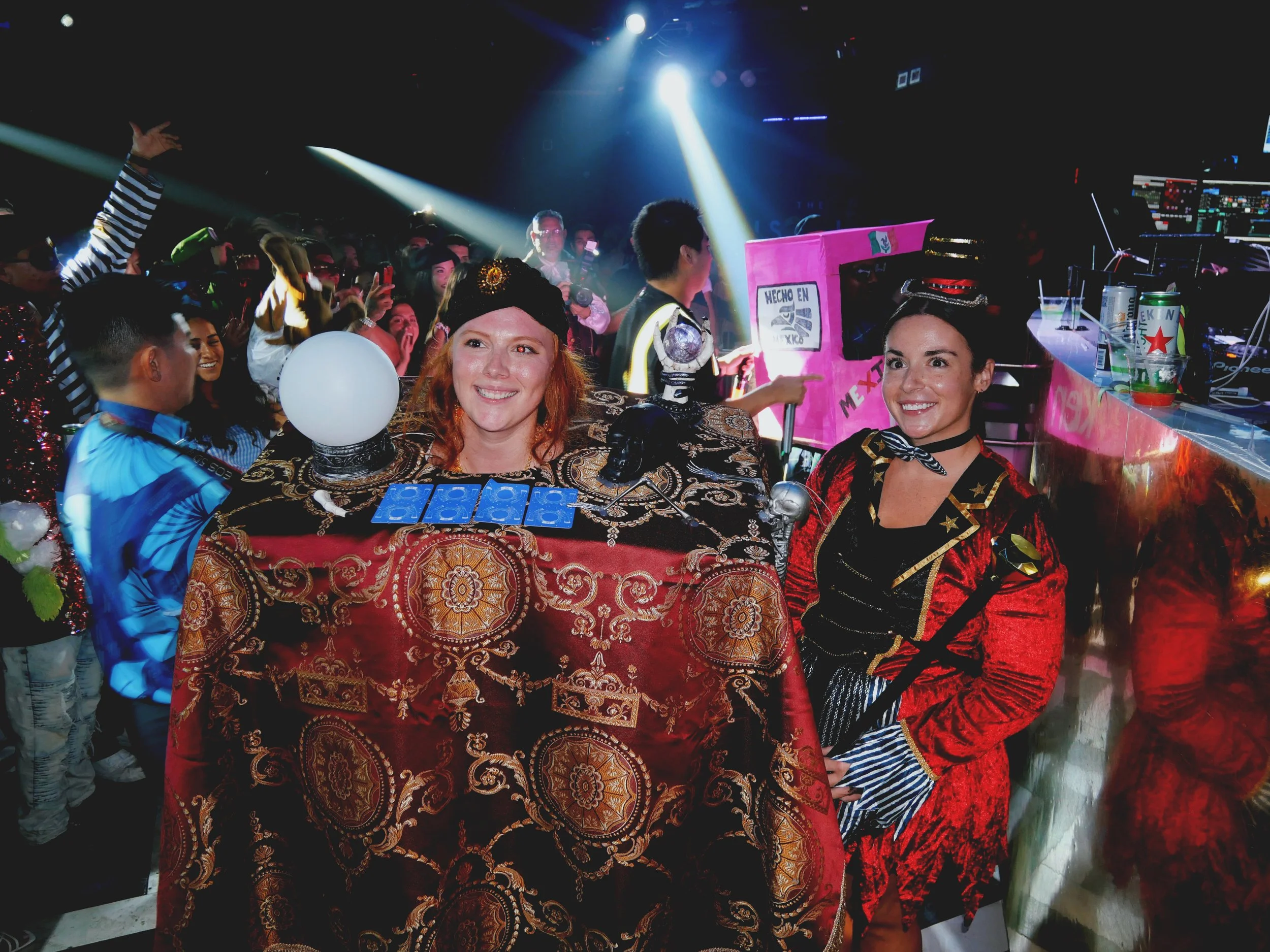 People dressed in colorful costumes and uniforms standing around a decorated table at a lively indoor event with party lights and a DJ booth.