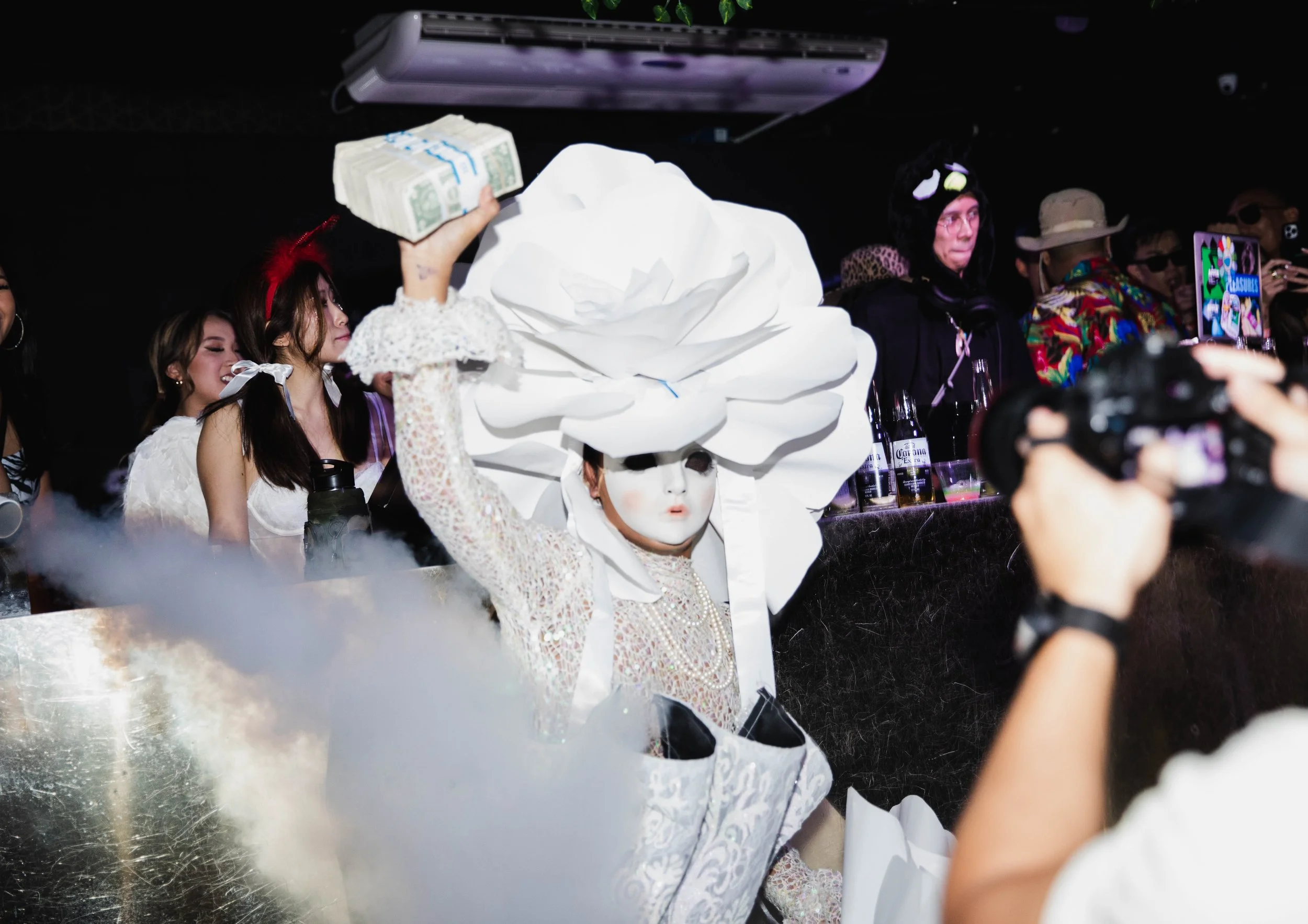 A person in a white lace outfit and theatrical mask, with a large white floral headpiece, is holding a stack of cash in a nightclub or party setting with other party-goers in the background.