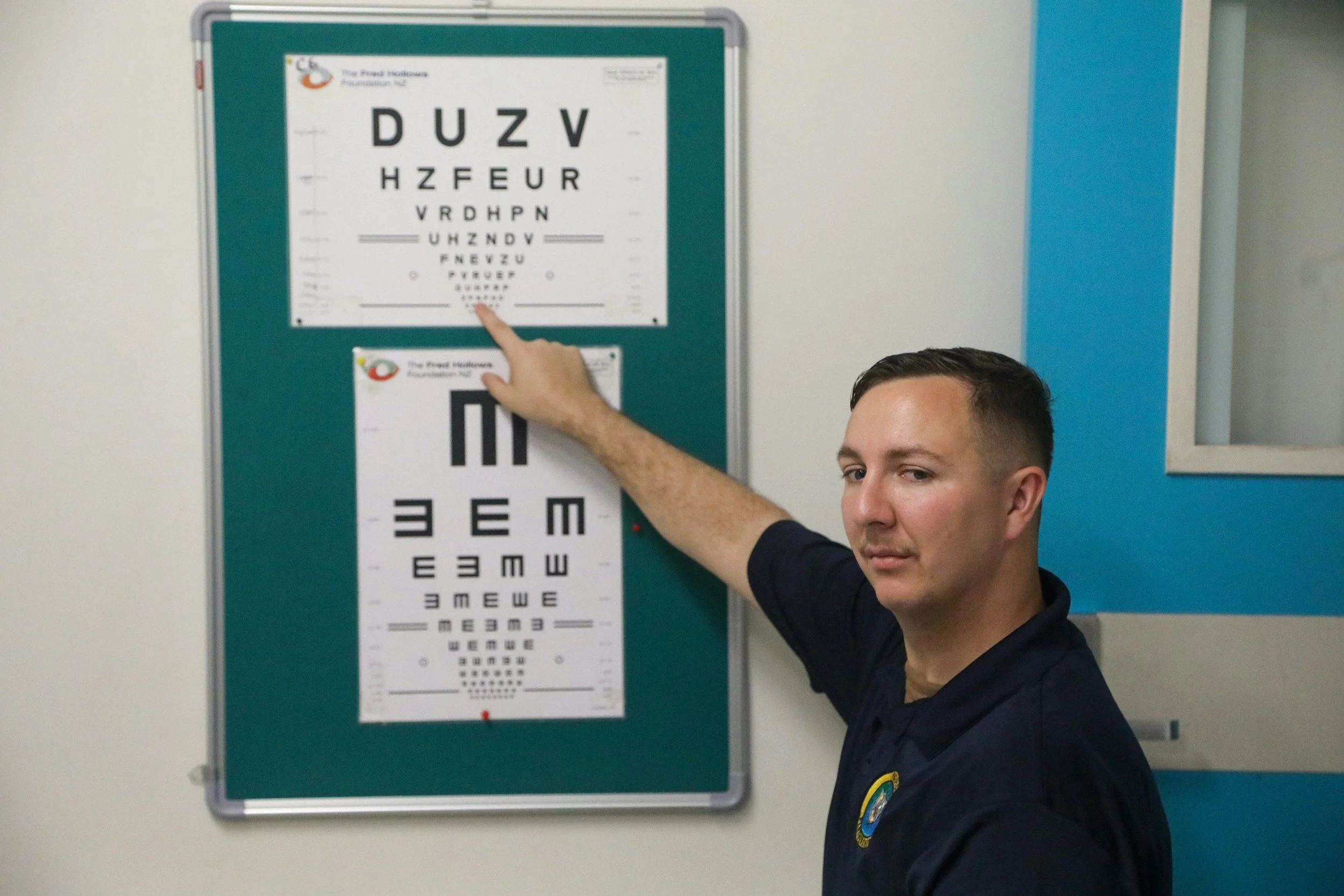 A young man with short dark hair is pointing at an eye chart on a green bulletin board. The eye chart has rows of black letters in a decreasing size, starting with large 'D U Z V' at the top and ending with smaller letters at the bottom. The young man is wearing a dark school uniform with a badge on the sleeve. The background includes a wall with a blue stripe and a window.