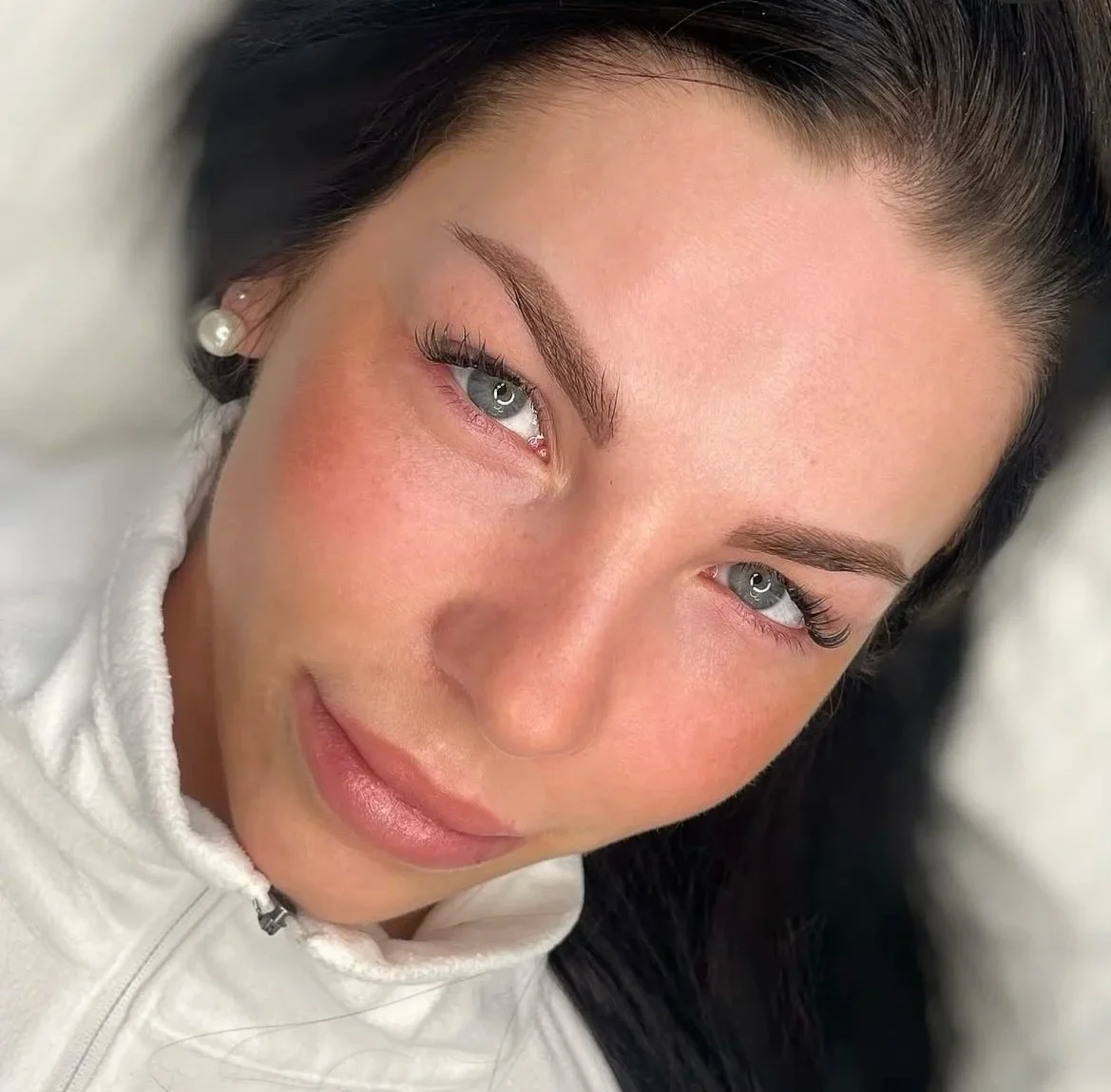 Nahaufnahme eines lachenden Mädchens mit blauen Augen, dunklem Haar, Perlenohrringen und einem weißen Hoodie.
