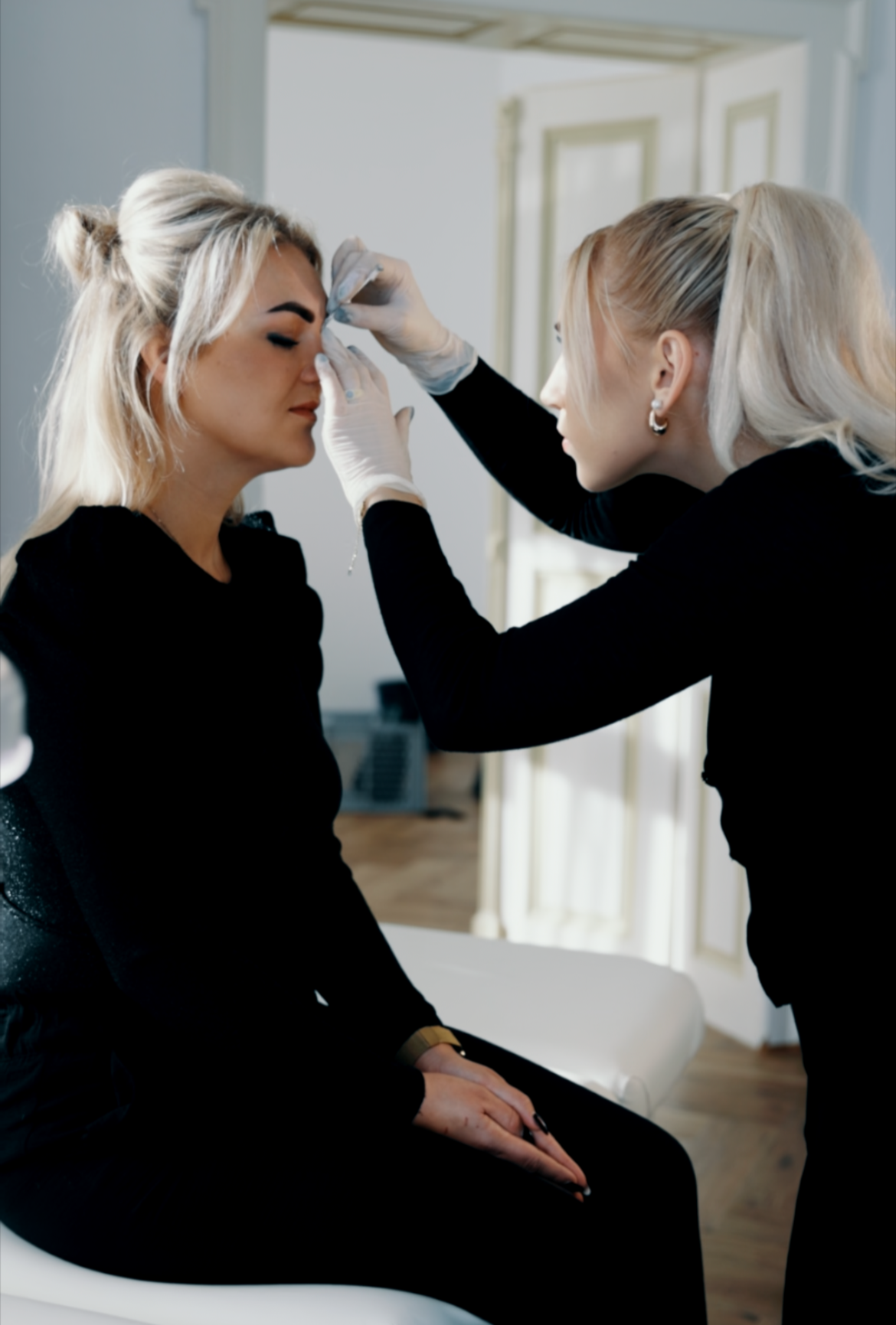 Zwei Frauen, eine sitzt mit geschlossener Augen und trägt schwarzes Kleid, die andere steht und trägt schwarze Kleidung, bereiten eine Körperkunst vor, indem sie an der Nase der sitzenden Frau arbeitet, in einem hellen Raum mit Holzfußboden.